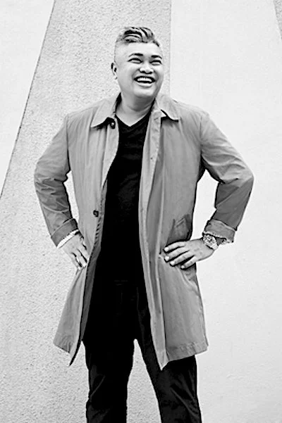 A person smiling outdoors, wearing a light jacket over a dark shirt, with hands on hips.