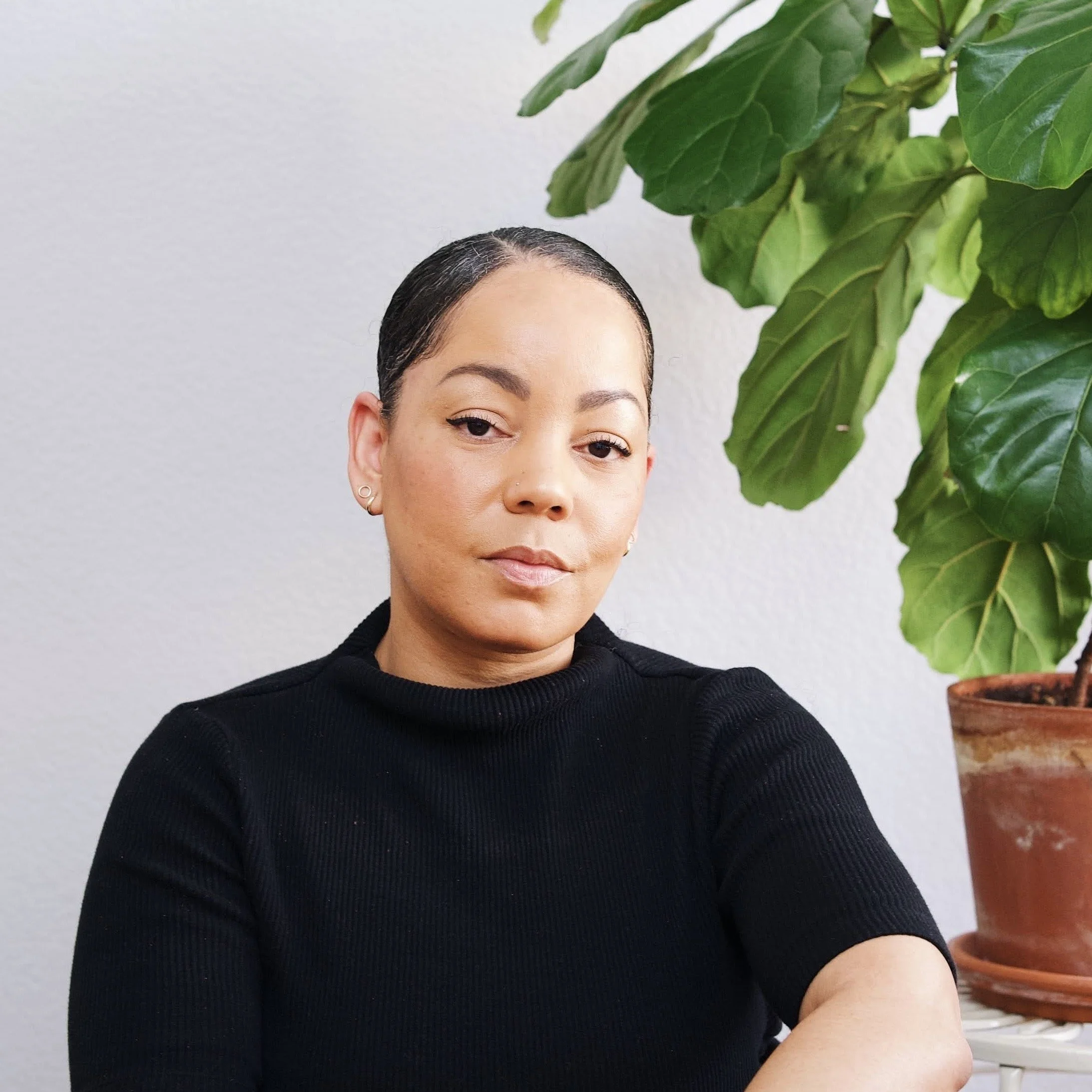 A woman with dark hair pulled back, wearing a black turtleneck, sitting next to a potted fiddle leaf fig plant with large green leaves, against a plain white wall.