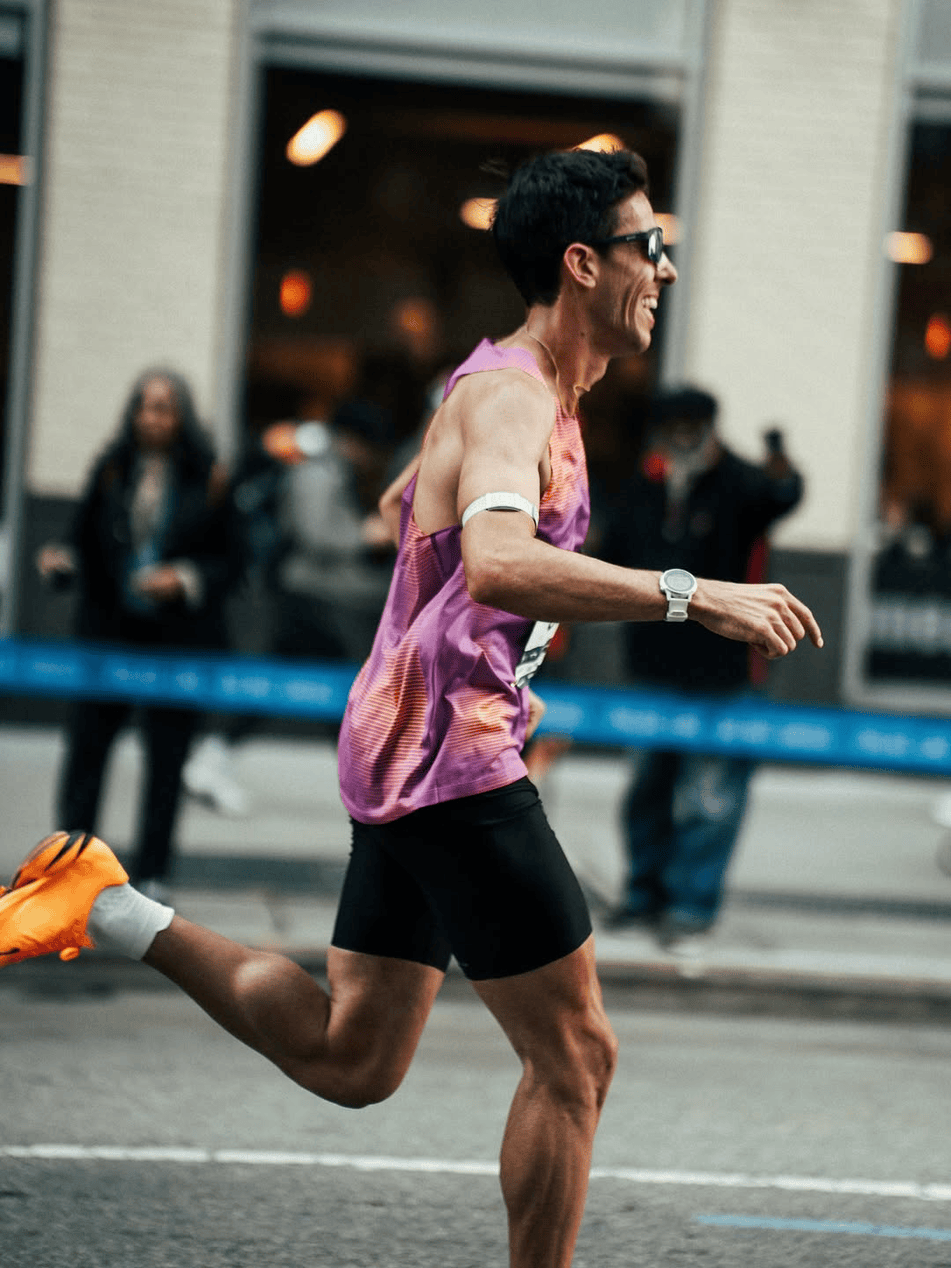 A smiling male runner in a purple tank top and black shorts participating in a race on a city street, with onlookers in the background.