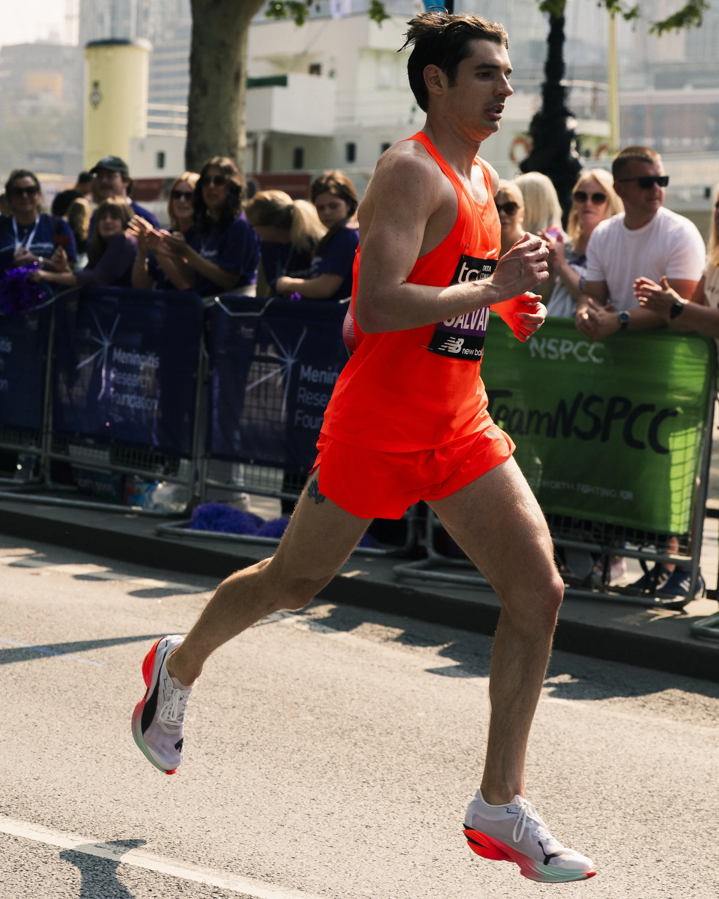 A male runner in orange athletic gear running in a race, with spectators clapping and cheering behind metal barricades.