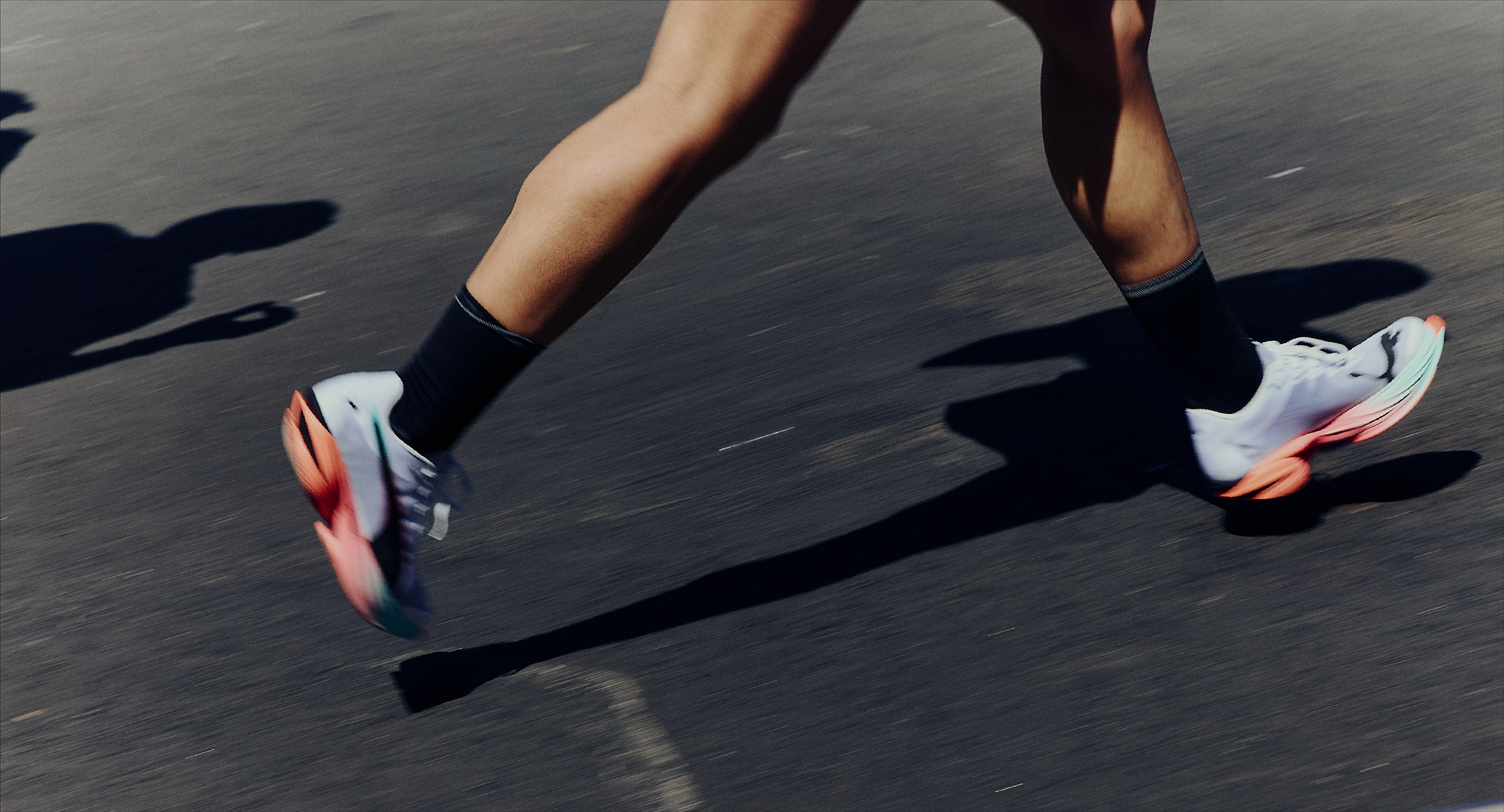 Close-up of a runner's legs and feet in motion on asphalt, wearing black socks and white running shoes with colorful soles.