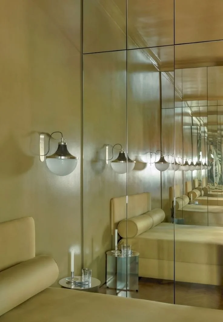 Chambre à coucher avec murs crème, appliques chromées aux murs, miroir reflétant la photo, table d'appoint métallique Hay