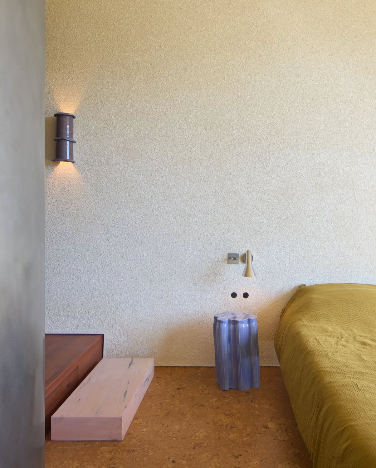 chambre avec sol liège par Bone Studio,  lit recouvert d'un drap moutarde, table basse bleue , applique arne jacobsen au lur, marches en marbre sur le côté gauche