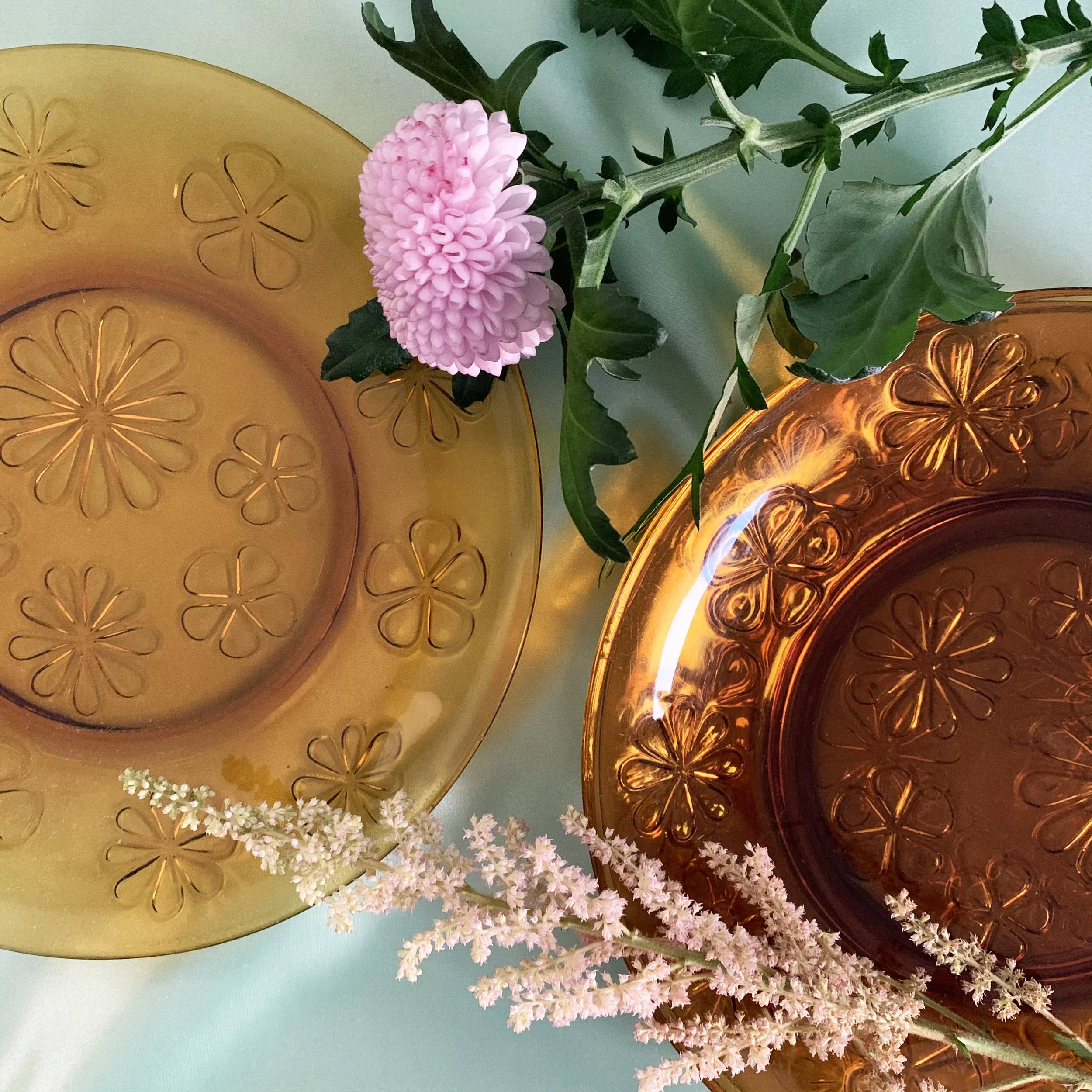 Deux plats en verre, un orange et un jaune, avec des motifs floraux, entourés de fleurs roses et blanches.