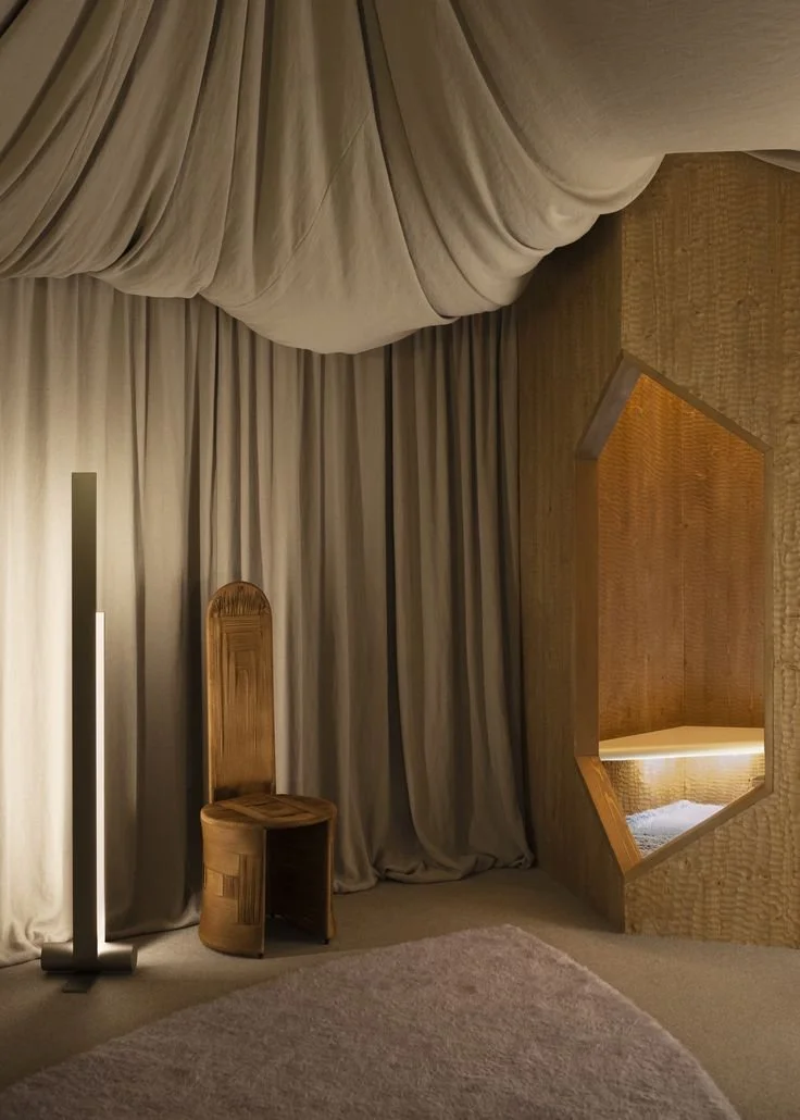 Pièce drapée avec rideaux beige, moquette sol, luminaire métallique, siège sculptural en bois