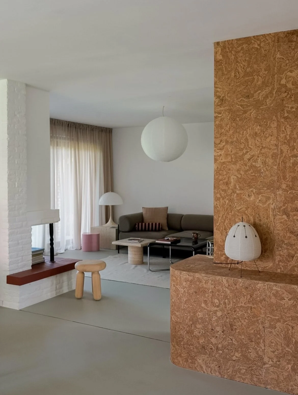 intérieur avec sol béton ciré, grande étagère plaquée liège avec lampe AKARI DESSUS, AU FOND UN CANAPÉ AVEC UNE LAMPE PANTHELLA SUR TABLE BASSE à coté et une fenêtre habillée de rideaux