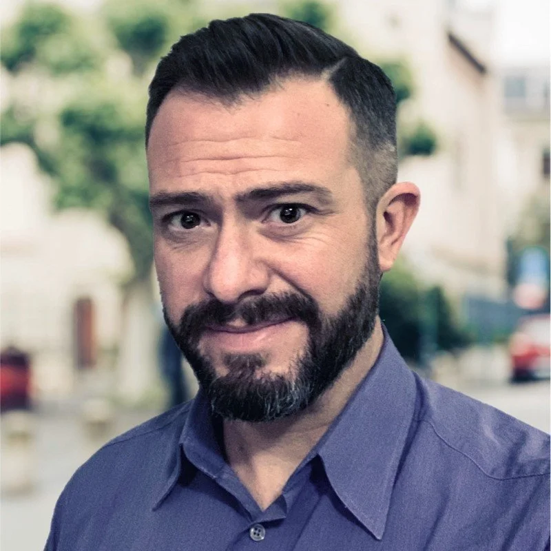 Portrait of a man with short dark hair and a neatly trimmed beard, wearing a blue button-up shirt. They are looking at the camera with a slight smile, standing outdoors with trees and buildings softly blurred in the background.