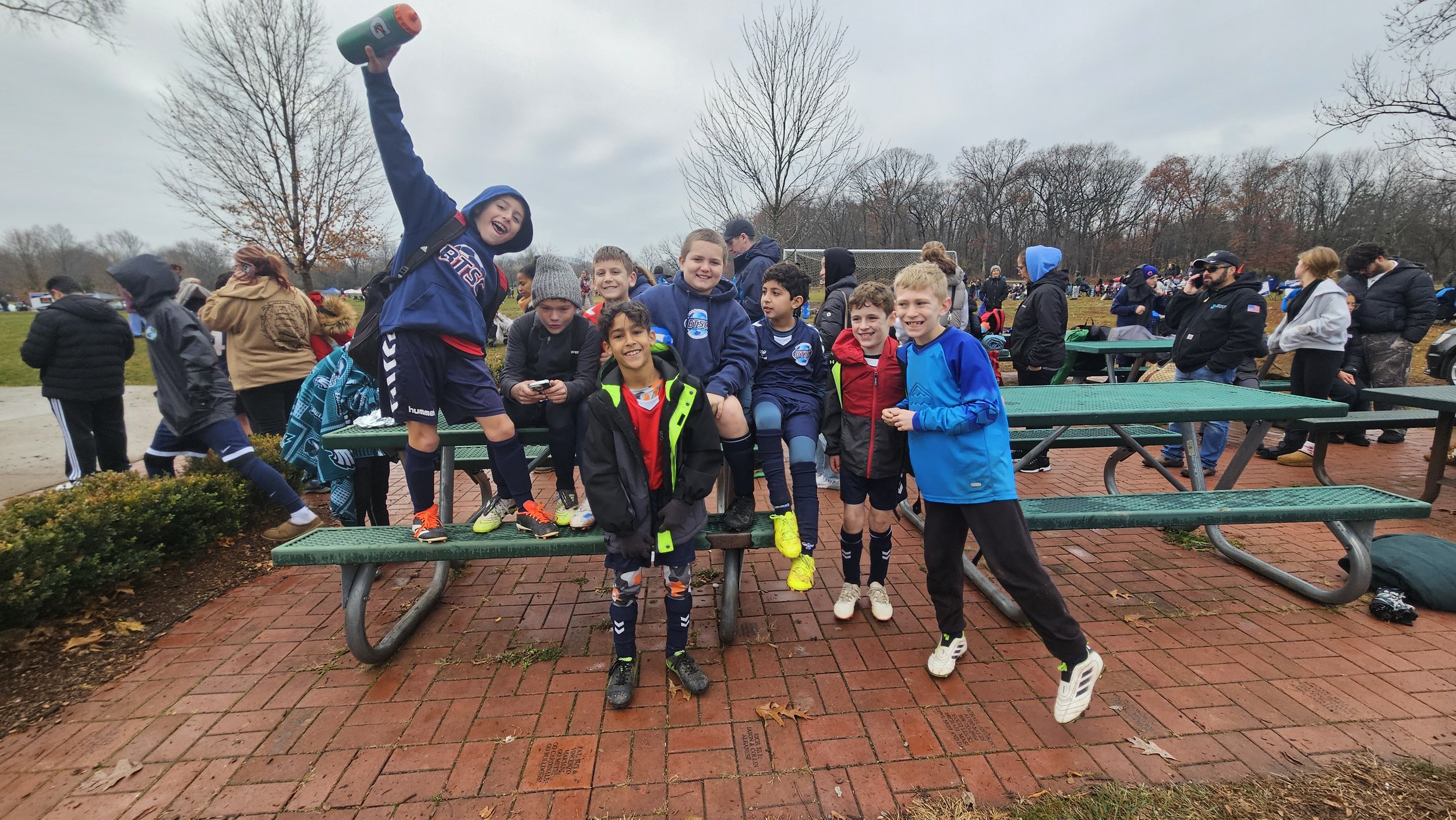 A group of children and teenagers gathering outdoors on a cloudy day, some wearing sports gear and jackets, with one boy raising his hand holding a water bottle and smiling. In the background, there are more people, a grassy field, trees with bare branches, and picnic tables.