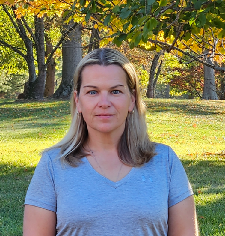 A woman with blonde hair wearing a light blue t-shirt standing outdoors in a park during fall with trees and grass in the background.