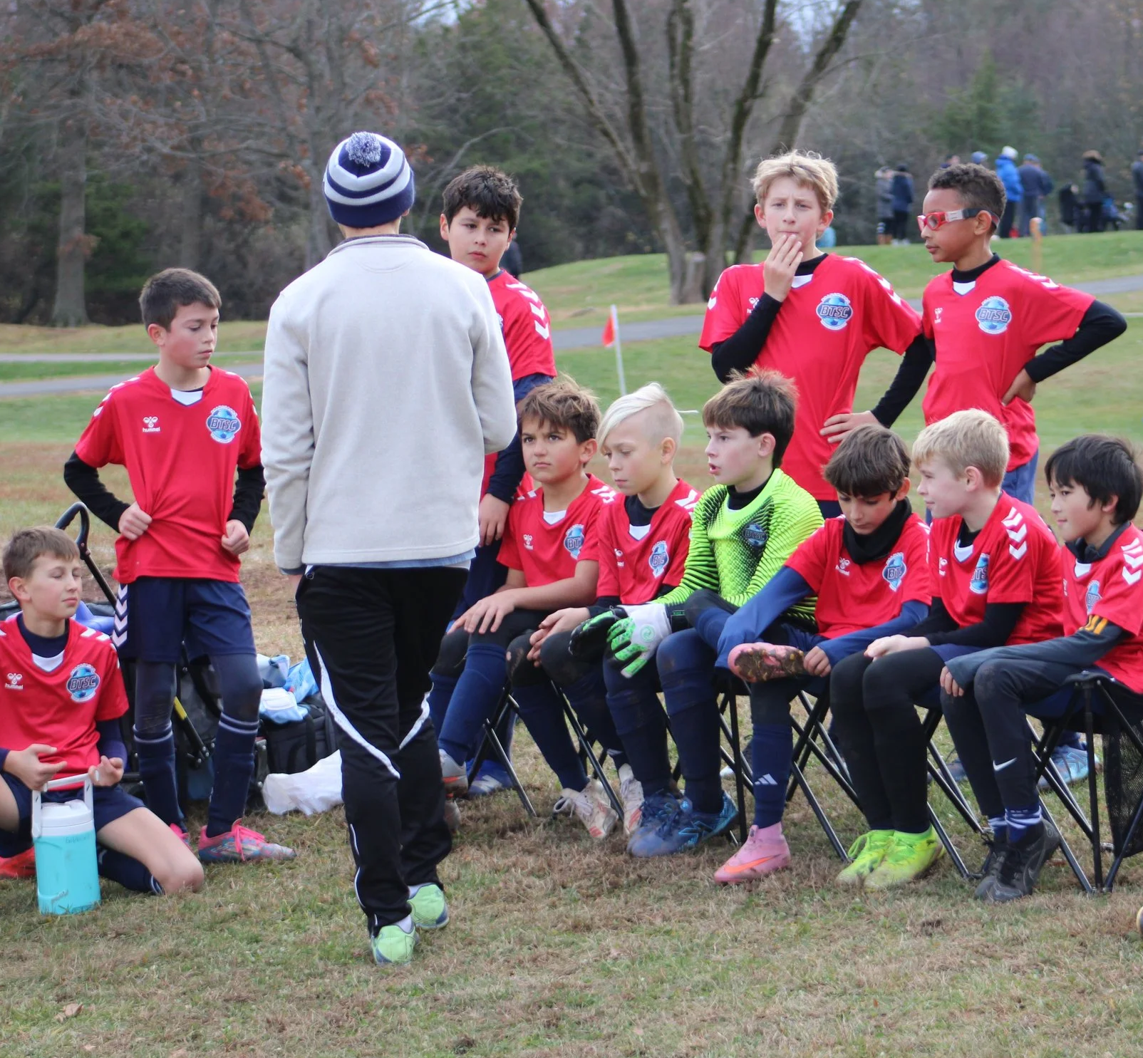 Youth soccer team listening to coach during a timeout on the field in a park with trees in the background.