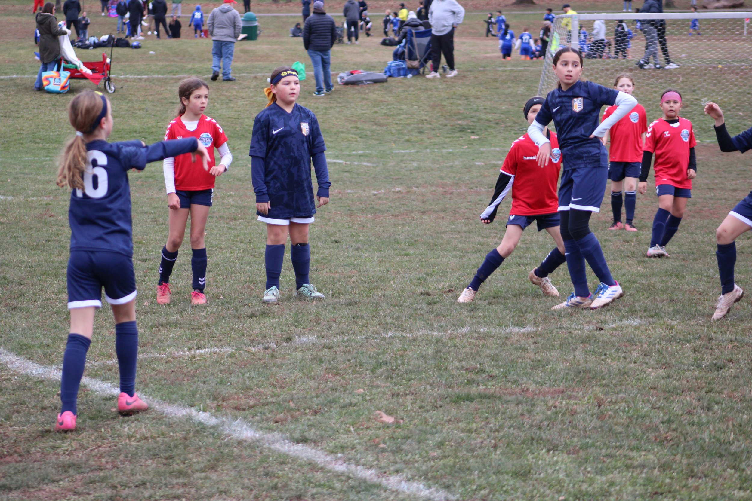 Children playing soccer on a grassy field during a youth sports event, with spectators and other players in the background.