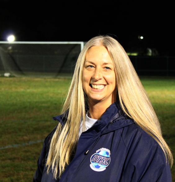 A smiling woman with long blonde hair wearing a navy blue jacket standing on a football field at night.
