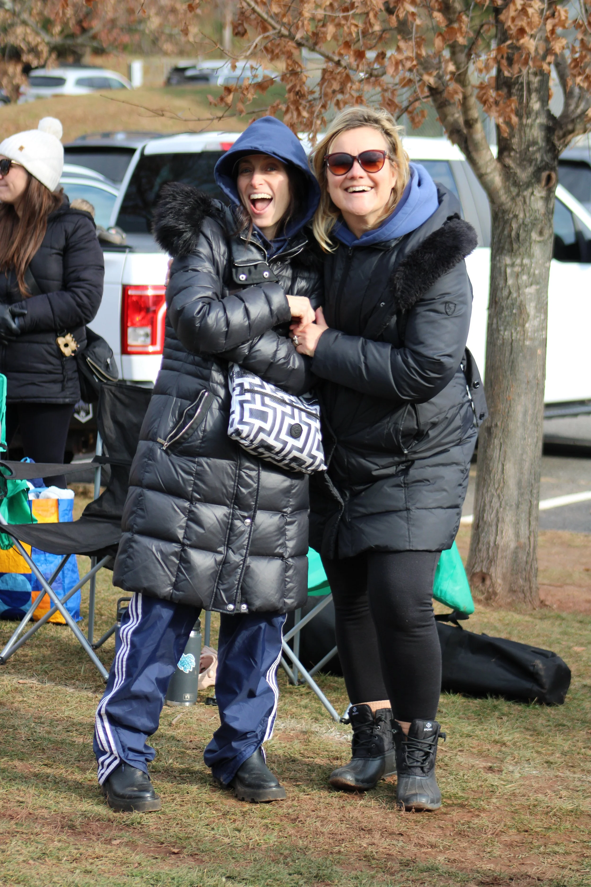 Two women smiling and holding hands outdoors on a fall day, dressed in warm clothing including black jackets, boots, and sunglasses, with a tree and parked cars in the background.