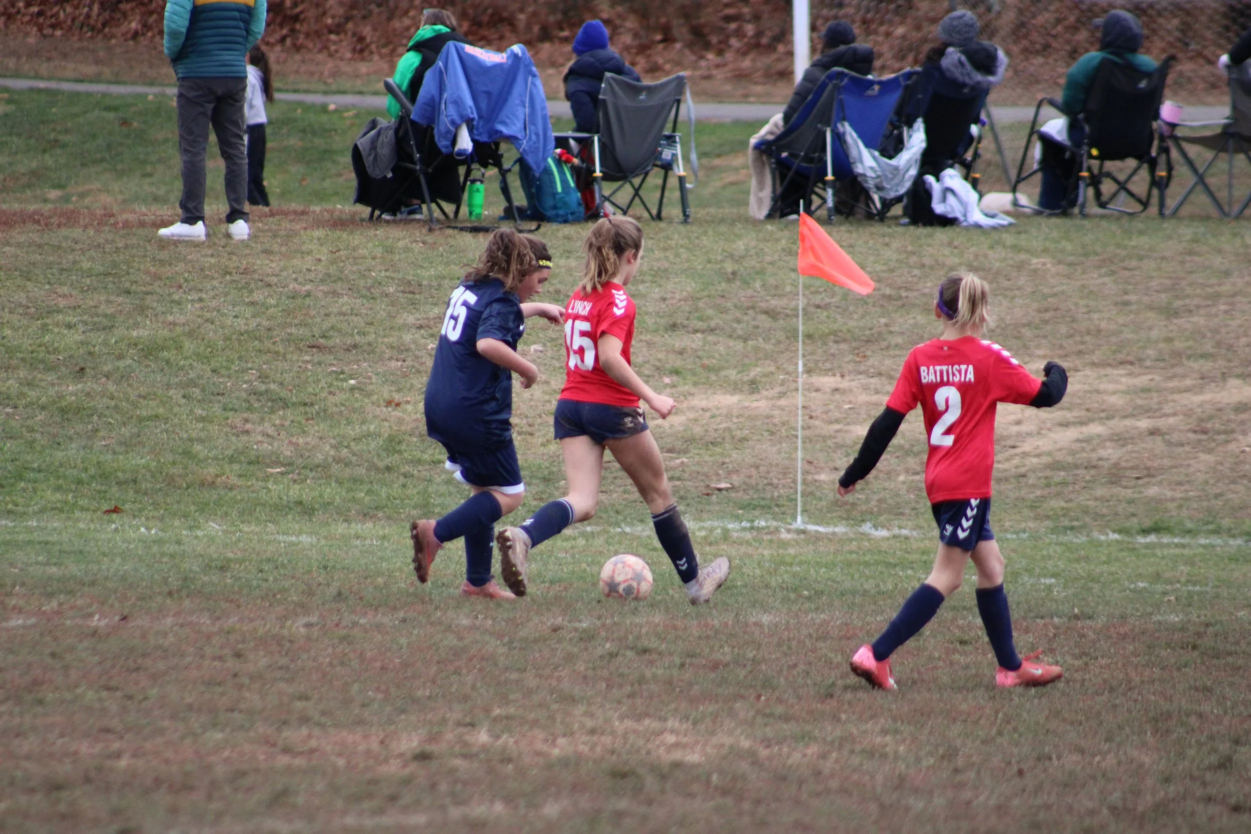 Young girls playing soccer on a grassy field, with players wearing red and navy uniforms, and spectators or players sitting on chairs in the background.