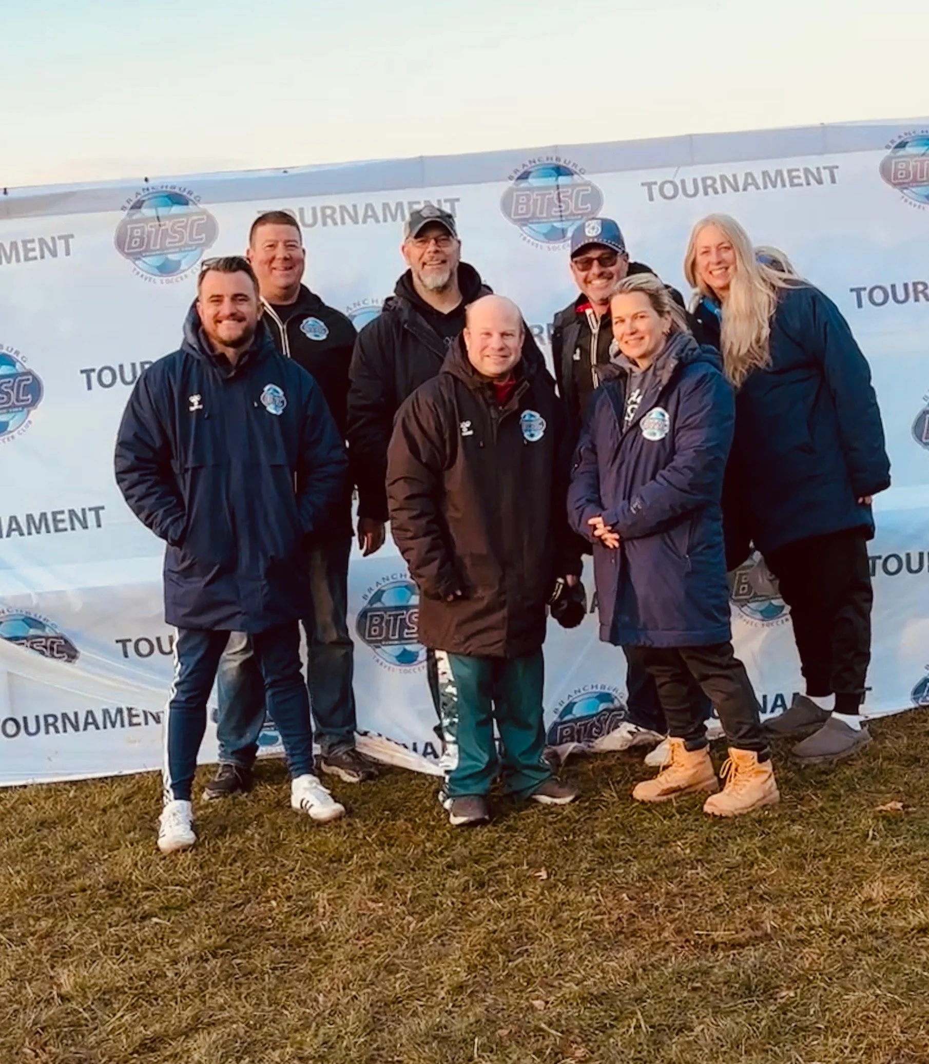 A group of seven people standing on muddy ground in front of a backdrop with the BTSC logo and the word "TOURNAMENT". They are dressed in outdoor jackets and smiling at the camera, with some wearing hats and sunglasses.