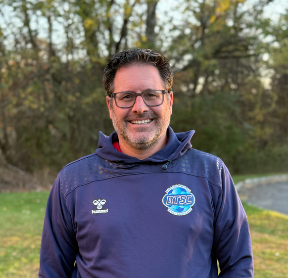 Man with glasses smiling outdoors wearing a navy blue jacket with logos, standing near a grassy area and trees.