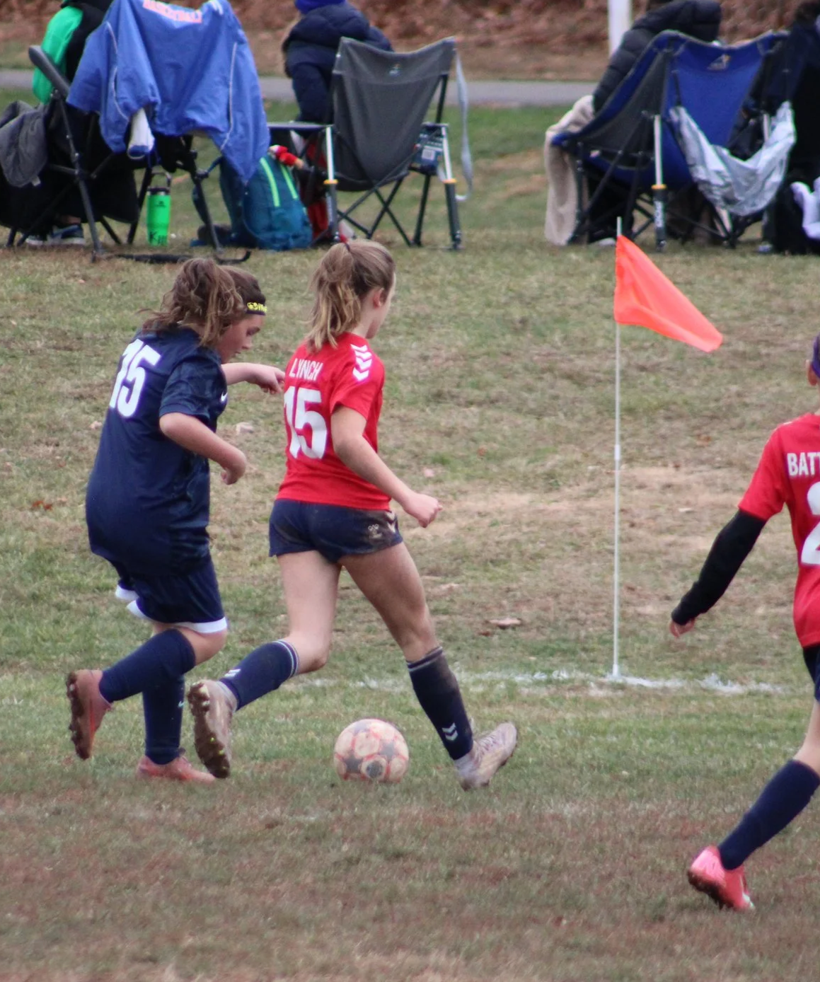 Children playing soccer on a field, with one girl in a red jersey and another girl in a navy jersey chasing the ball near a corner flag.