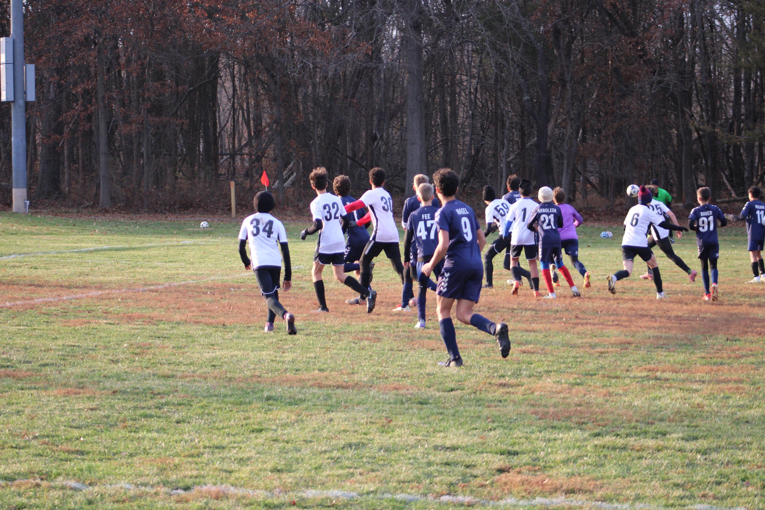 Group of children playing soccer on a grassy field in autumn, with a wooded area in the background.