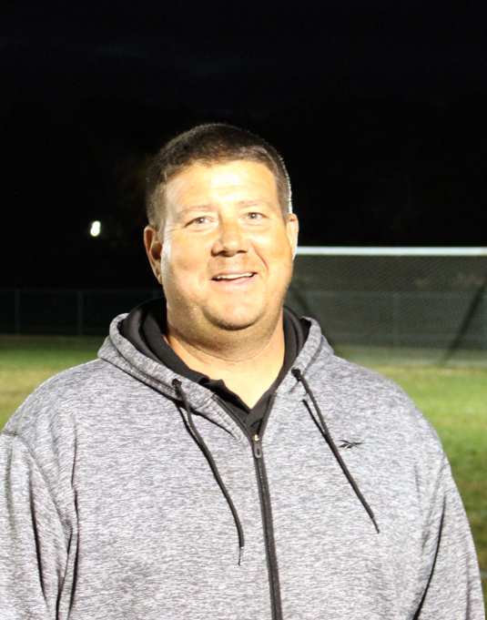 A man smiling outdoors at night, wearing a gray zip-up hoodie with a black shirt underneath, on a field with a fence and dark sky in the background.