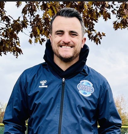 Man smiling outdoors, wearing a blue jacket with a BTSC logo, standing under a tree with autumn leaves.
