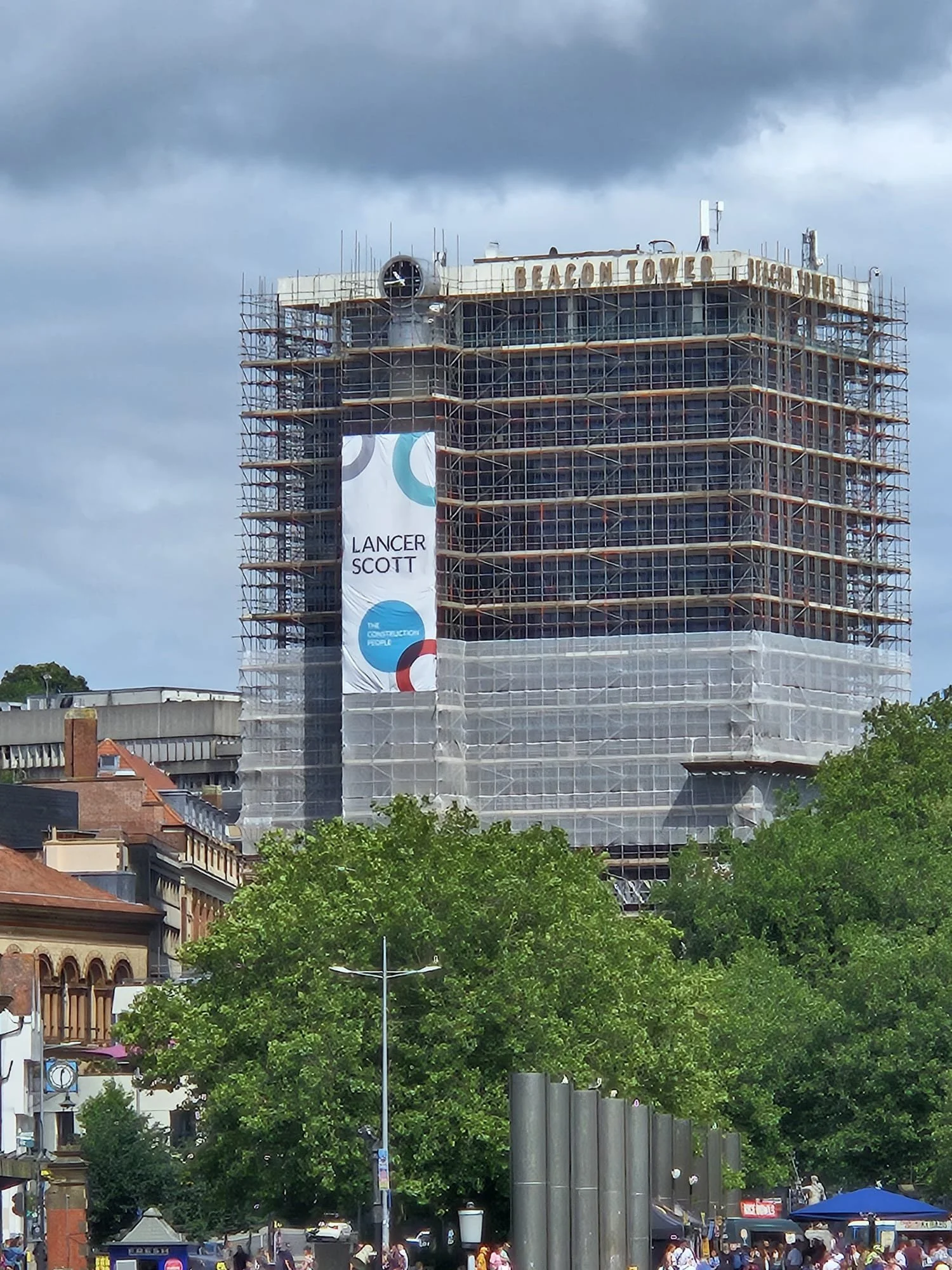 A tall building under renovation with scaffolding, and a banner reading 'Lancer Scott'. Trees and people at street level in the foreground.