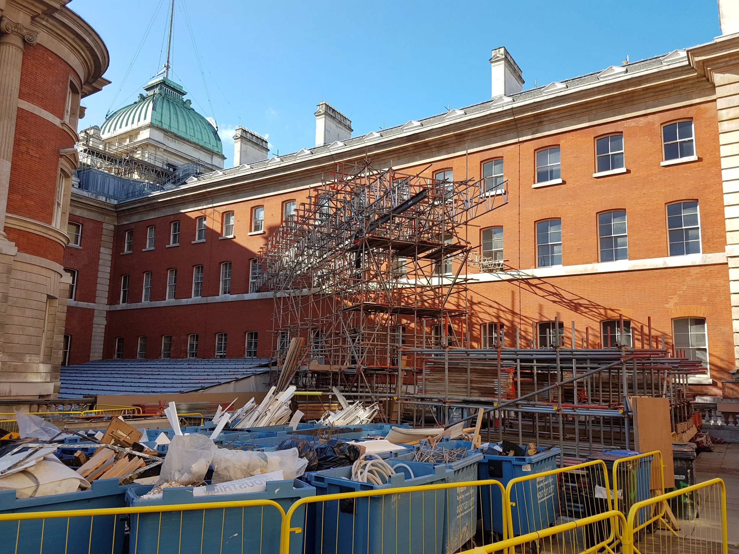 Construction site with scaffolding and building materials in front of a brick building with multiple windows and a domed roof in the background.