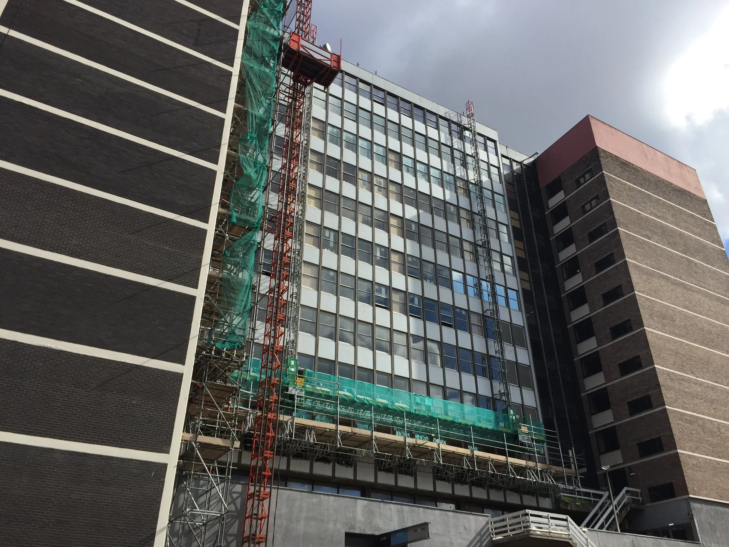 A multi-story building under construction with scaffolding, a red tower crane, and glass windows, set against a cloudy sky.