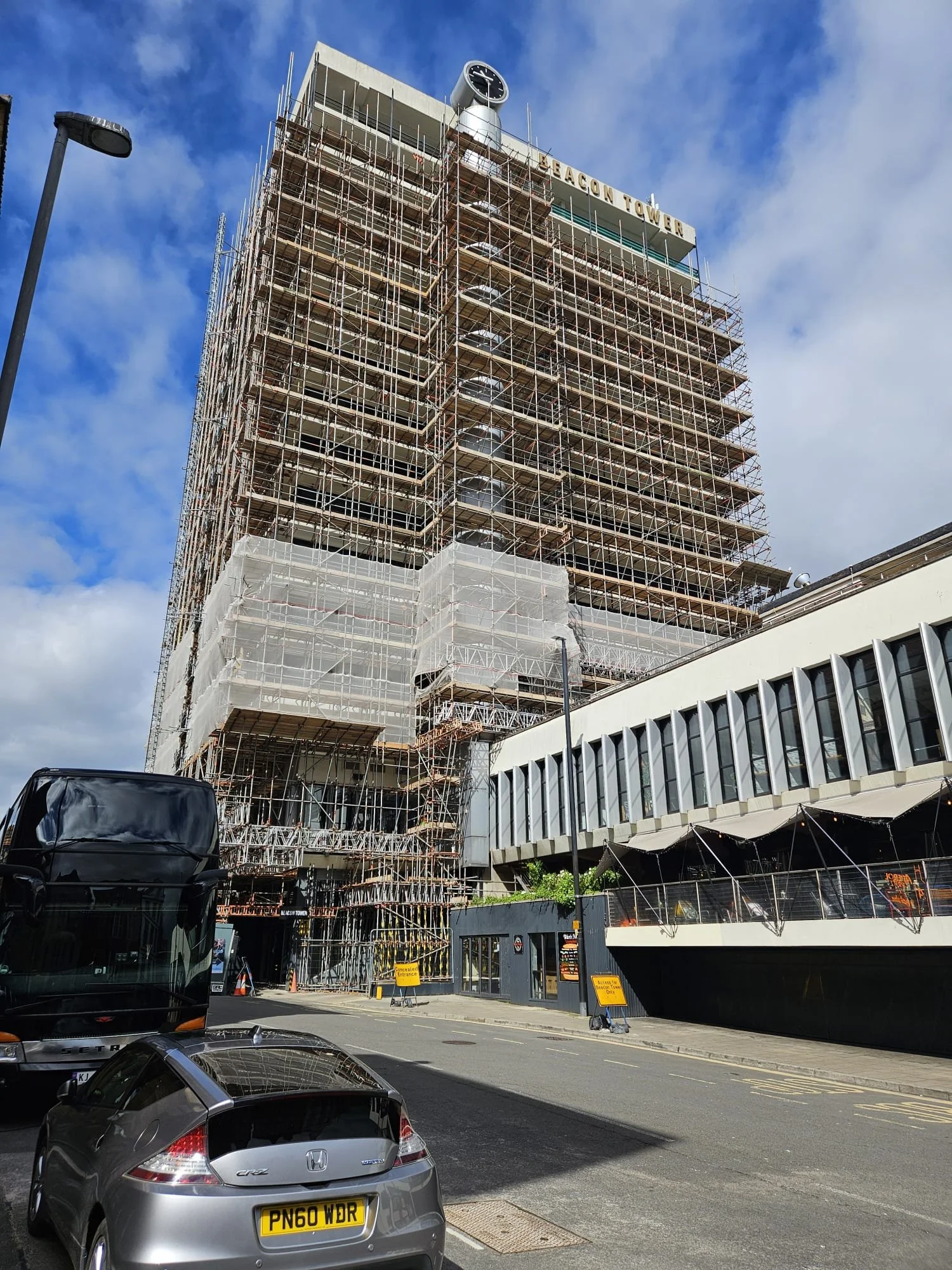 High-rise building under construction with scaffolding surrounding it, labeled 'Beacon Tower,' parked cars and a bus nearby, partly cloudy sky.