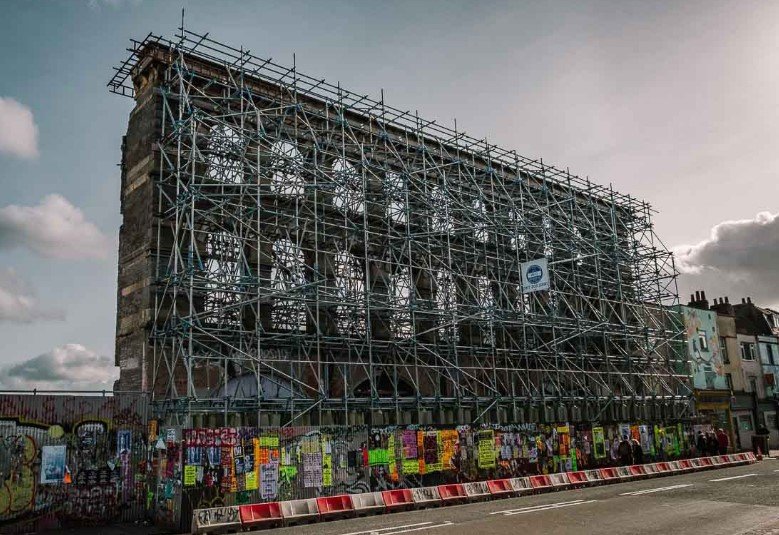 A building covered in scaffolding with graffiti and posters at the base, under a cloudy sky.