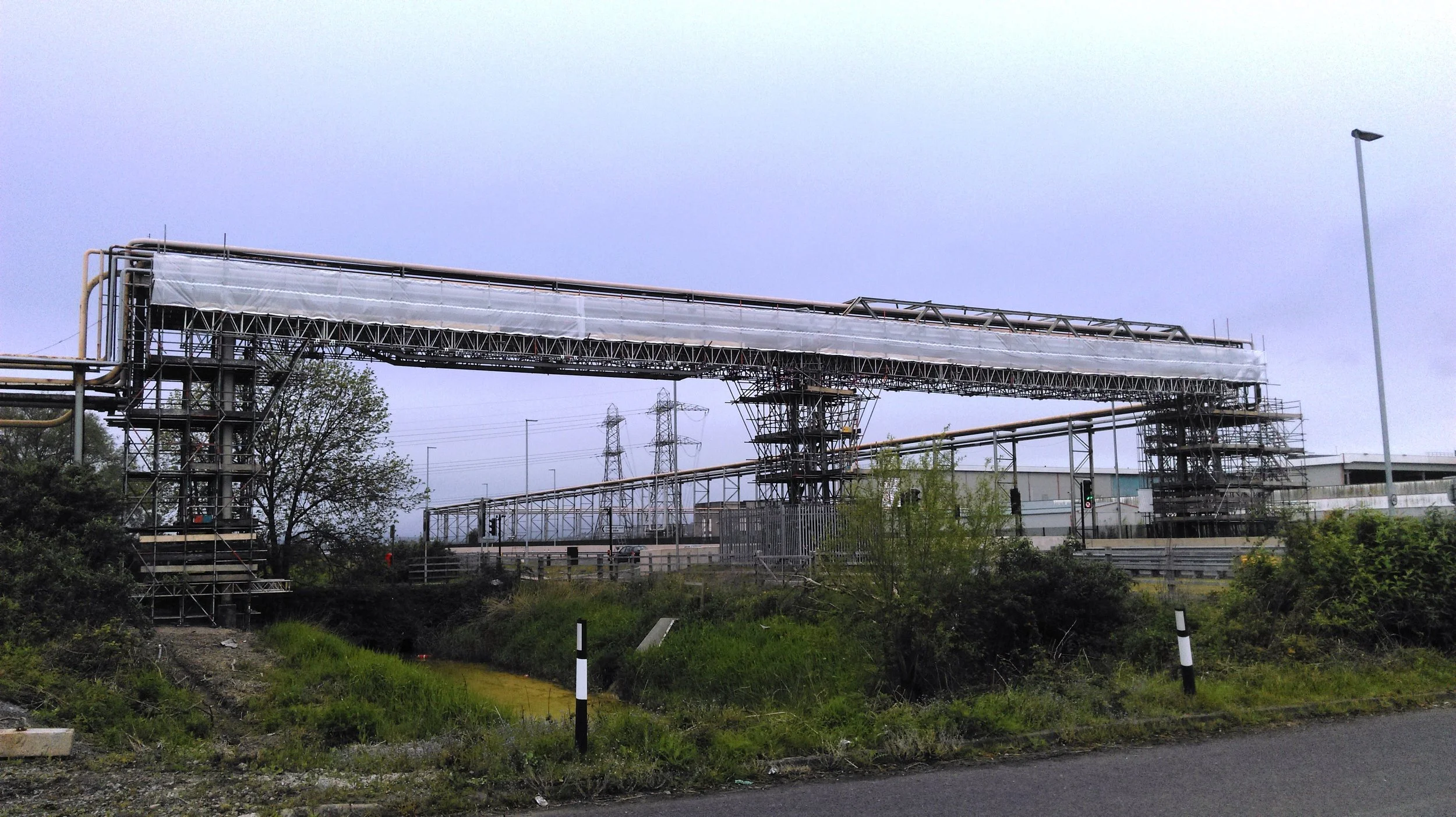 Industrial pipeline system under construction with scaffolding and a streetlight on a cloudy day.