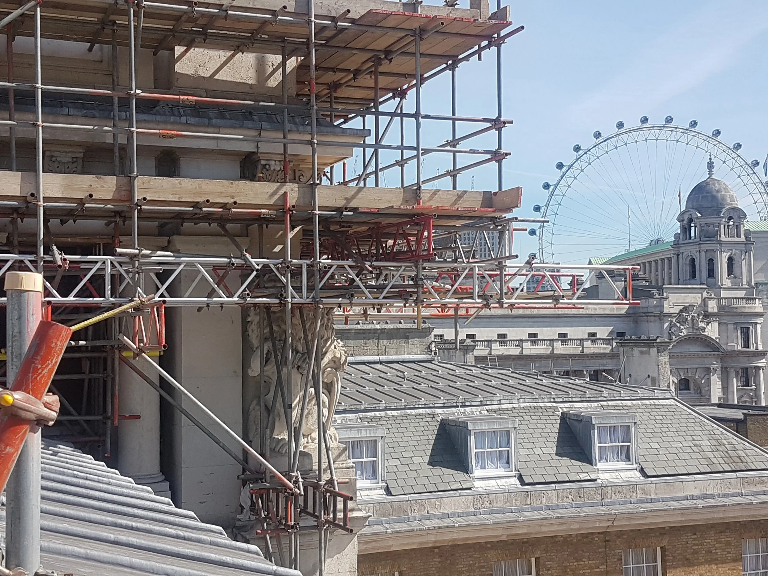 Construction scaffolding around a historic building with detailed stone carvings, and a large Ferris wheel visible in the background.