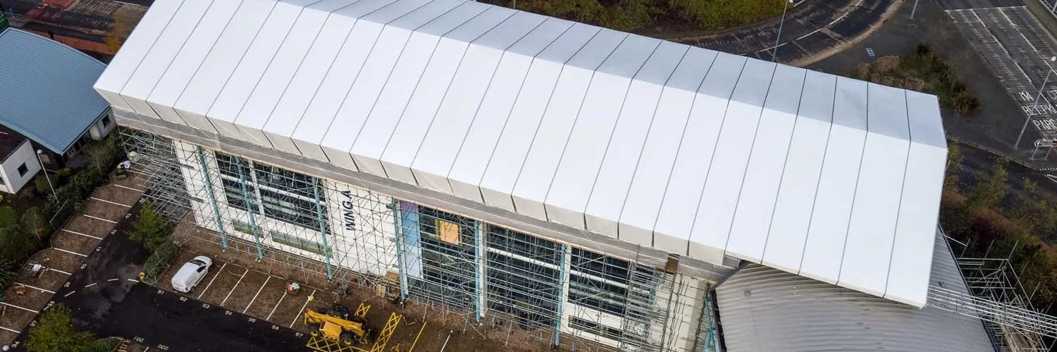 Aerial view of a modern building under construction with scaffolding on the facade, parking lot with few cars, and a road nearby.