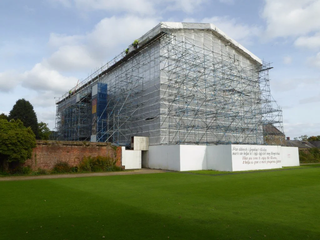 A building covered in scaffolding and protective sheeting under construction, with a white wall at its base and a green grassy area in the foreground, under a partly cloudy sky.