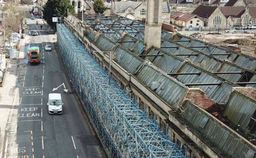 View from above of a street with a large construction scaffolding structure along the side of a building, which has a damaged roof with toppled panels.