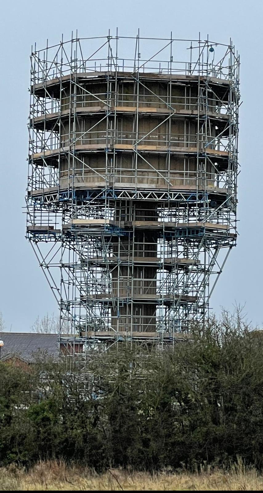 A tall, cylindrical water tower surrounded by metal scaffolding under construction, with trees and buildings in the background.