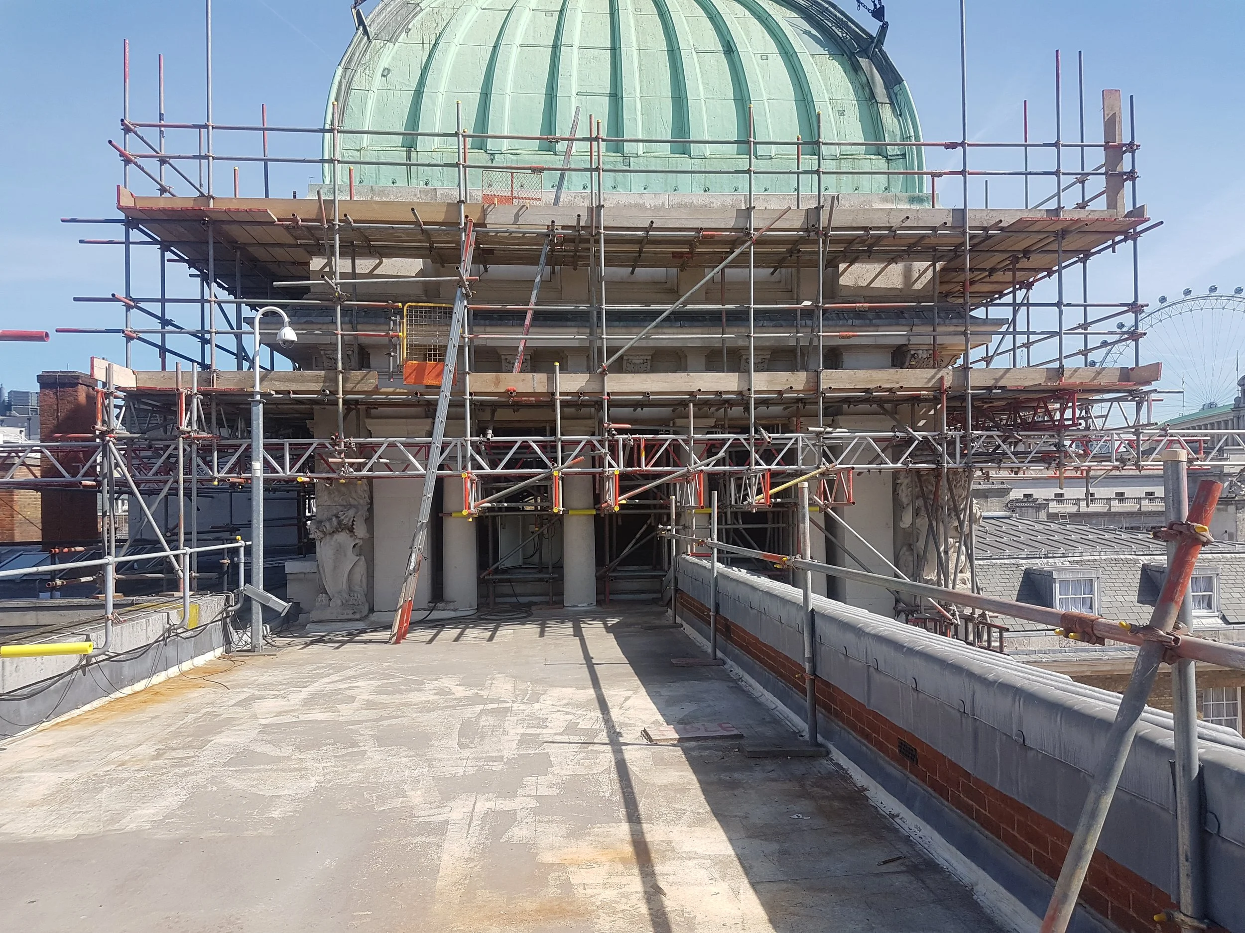 Scaffolding surrounding a historic building with a green dome under renovation on a sunny day.