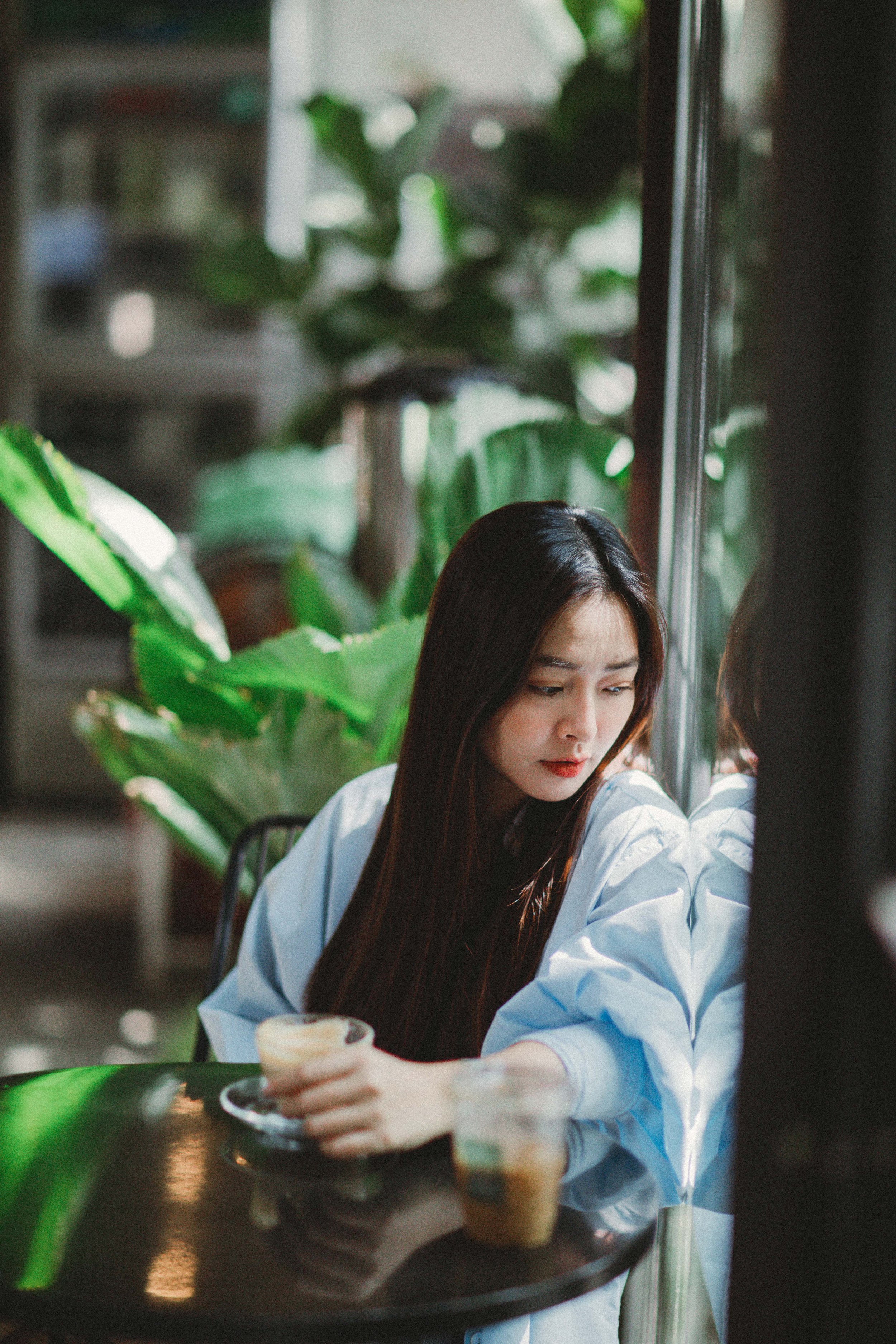 A young woman with long dark hair, wearing a light blue jacket, sitting at a table with a tray holding two cups of coffee or tea, looking out a window in a cafe with lush green plants in the background.