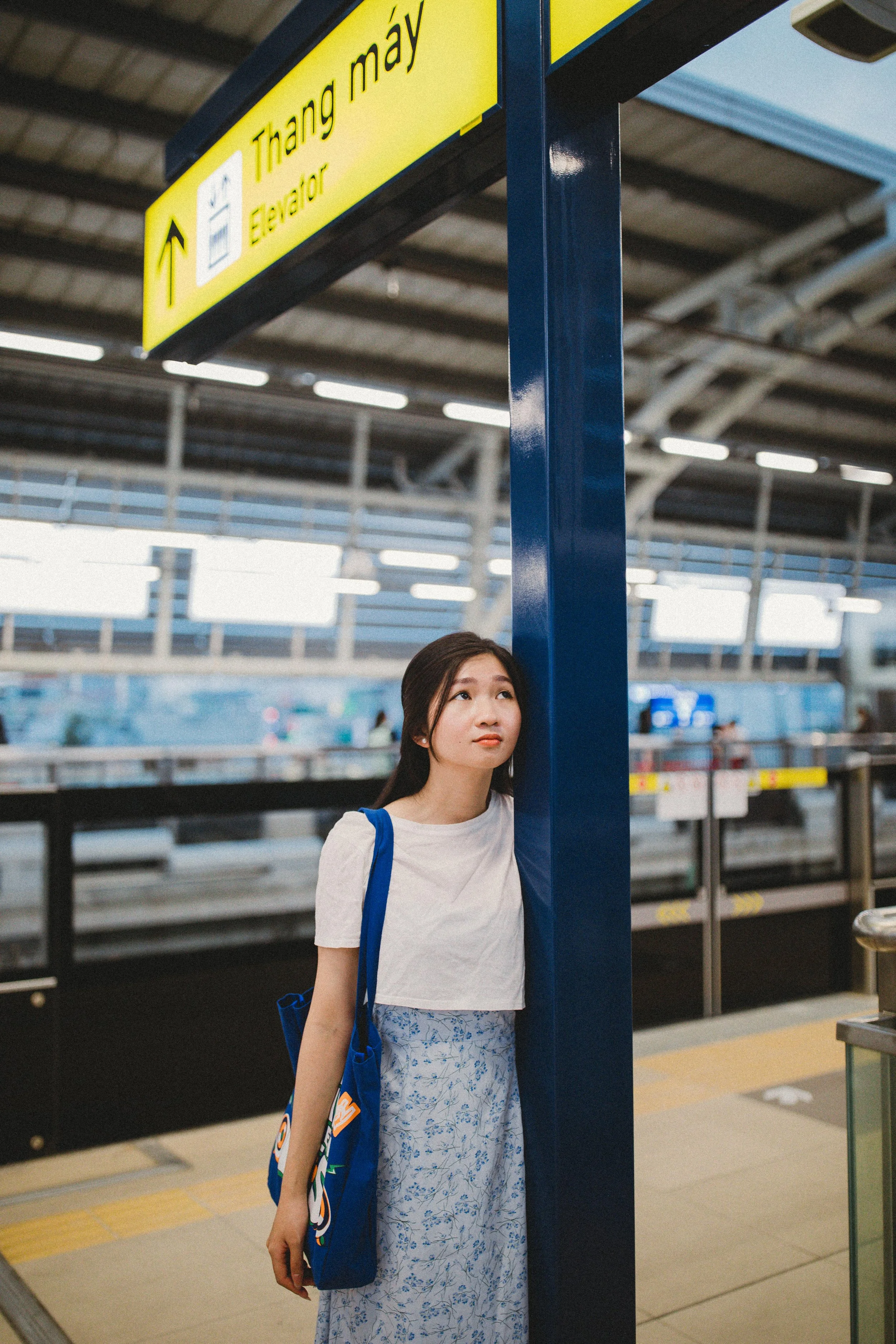 Young woman standing at a train station platform, leaning against a blue pole, looking thoughtful, with a yellow sign overhead indicating directions to Thang May elevator.