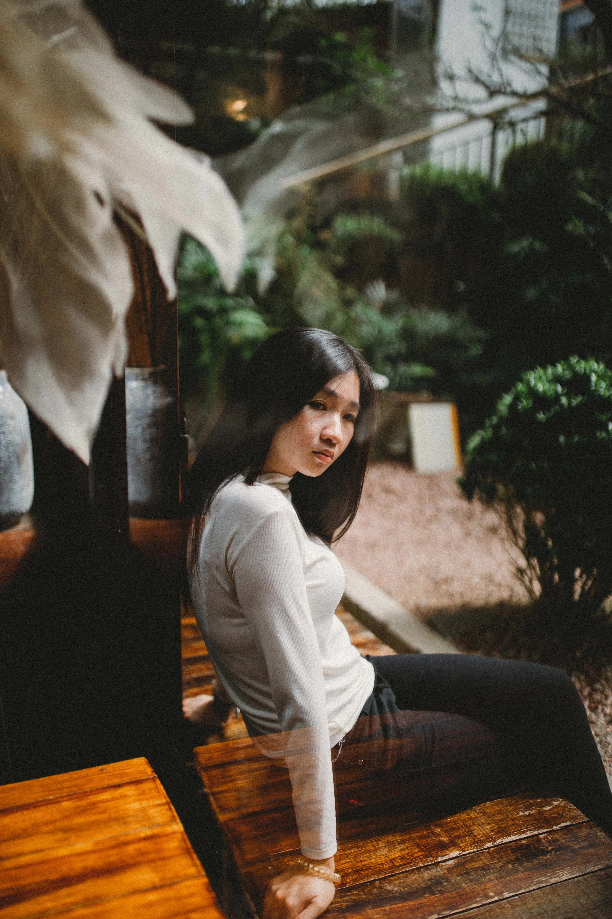 A woman with shoulder-length dark hair sitting on a wooden bench outdoors, surrounded by green plants and trees, looking at the camera.