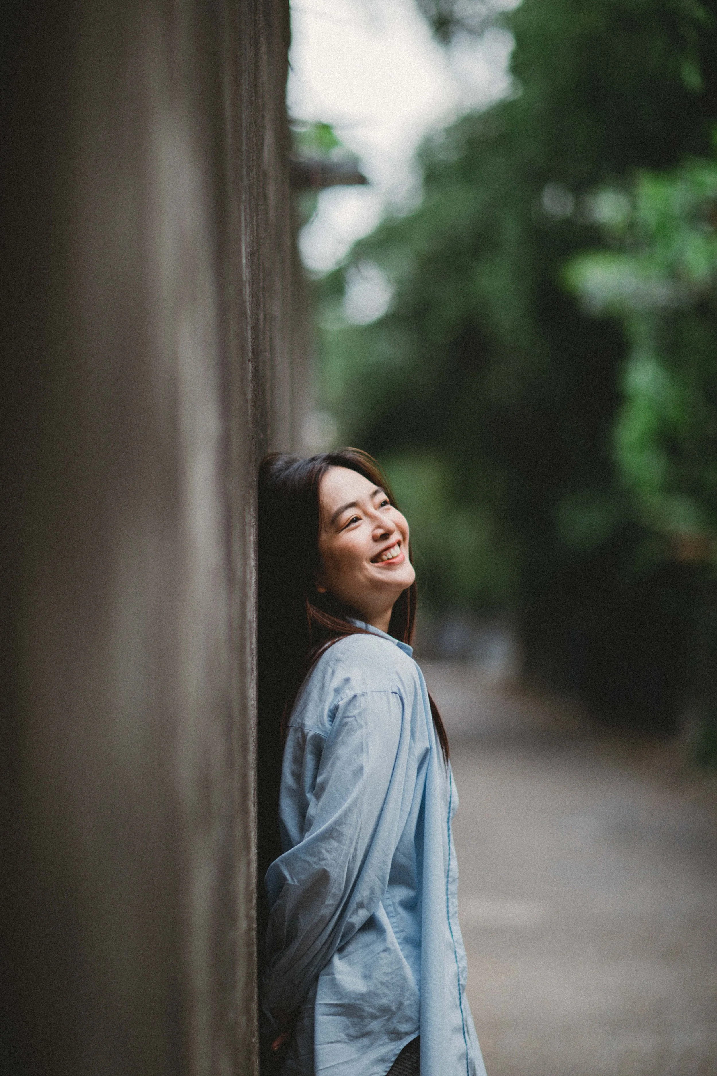 A woman with long hair leaning against a wall outdoors, smiling and looking to the side, with trees in the background.