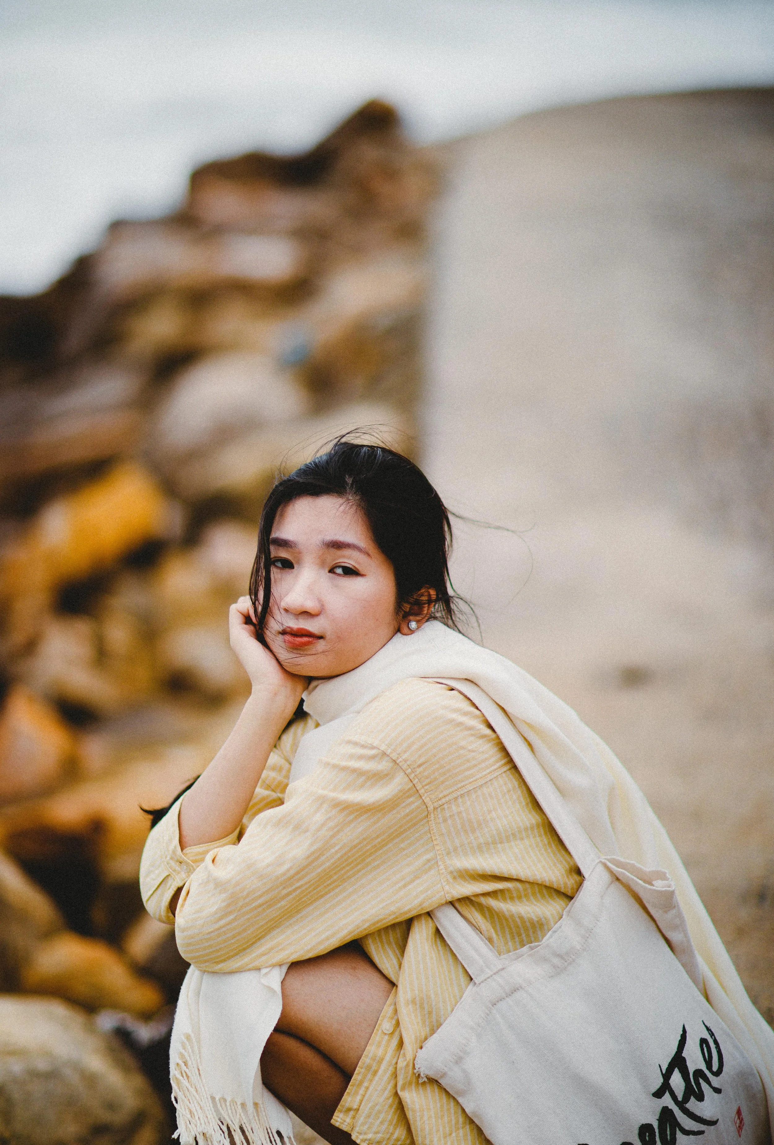 Young woman sitting outdoors near rocks, wearing a yellow striped shirt and carrying a beige tote bag, looking at the camera with a calm expression.