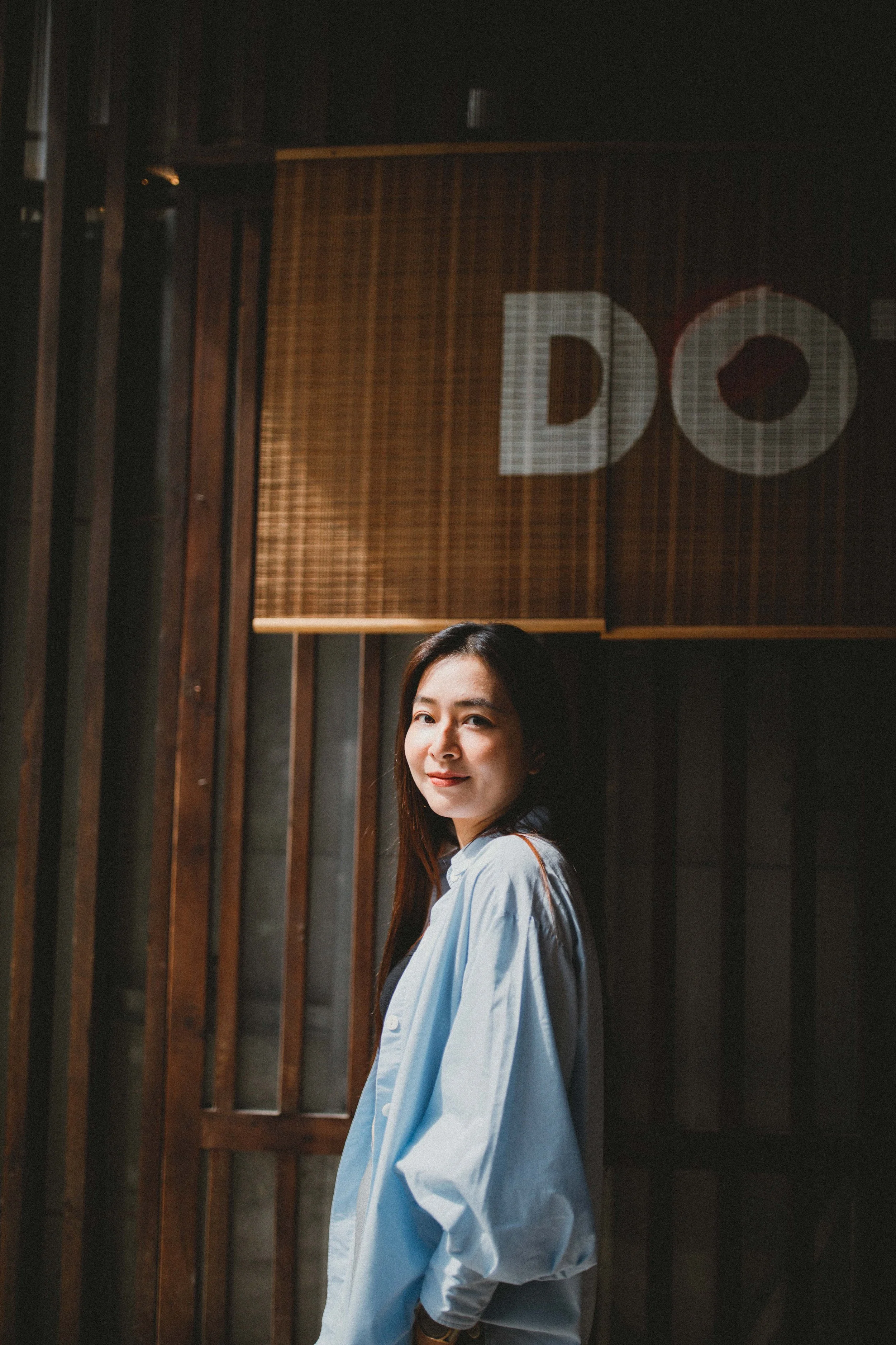 A woman with long dark hair wearing a light blue shirt, standing indoors in front of a wooden wall with vertical slats and a sign with the letters "DO".