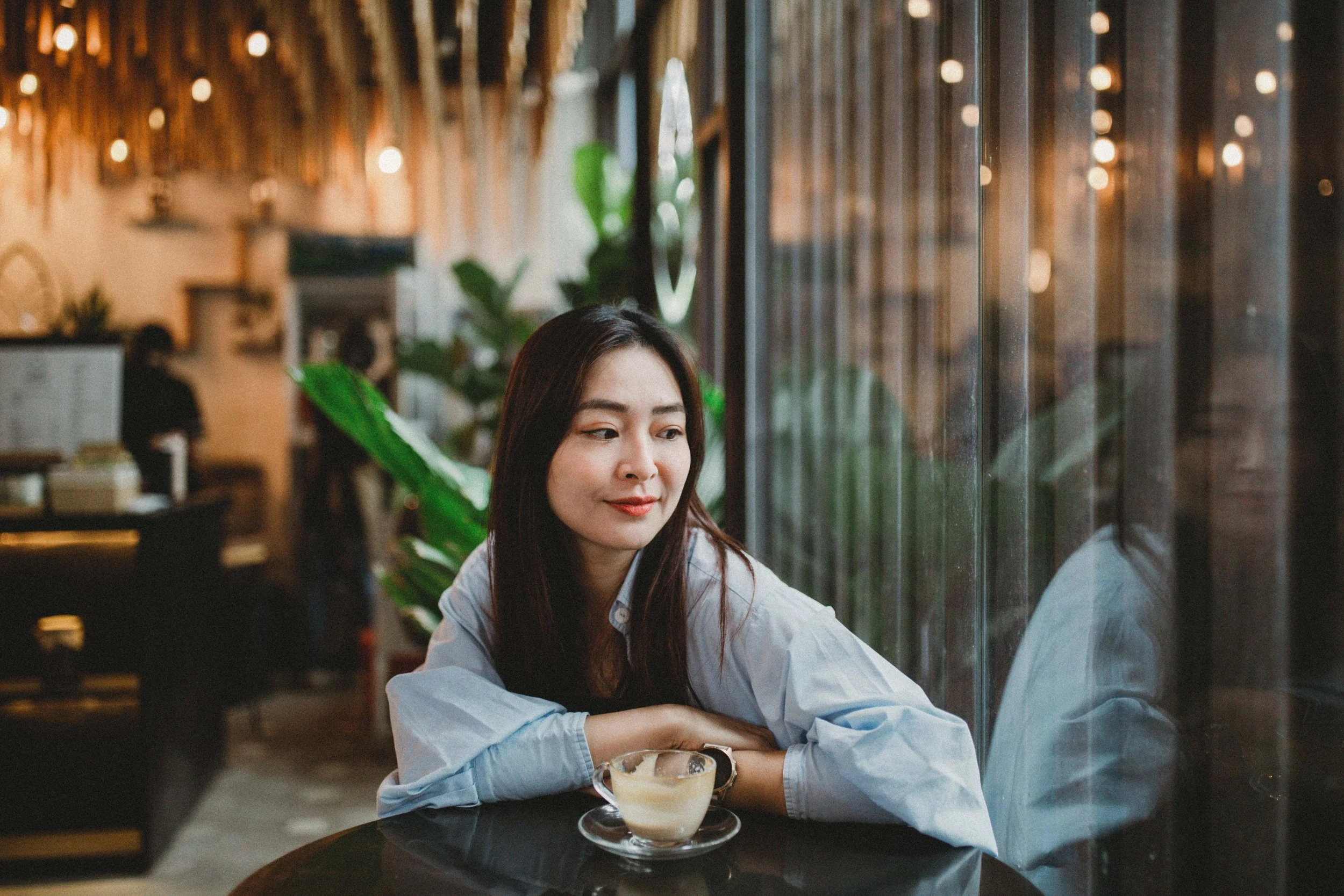 Young woman sitting at a coffee shop table, looking out the window with a cup of coffee or tea in front of her, surrounded by green plants and warm lighting.