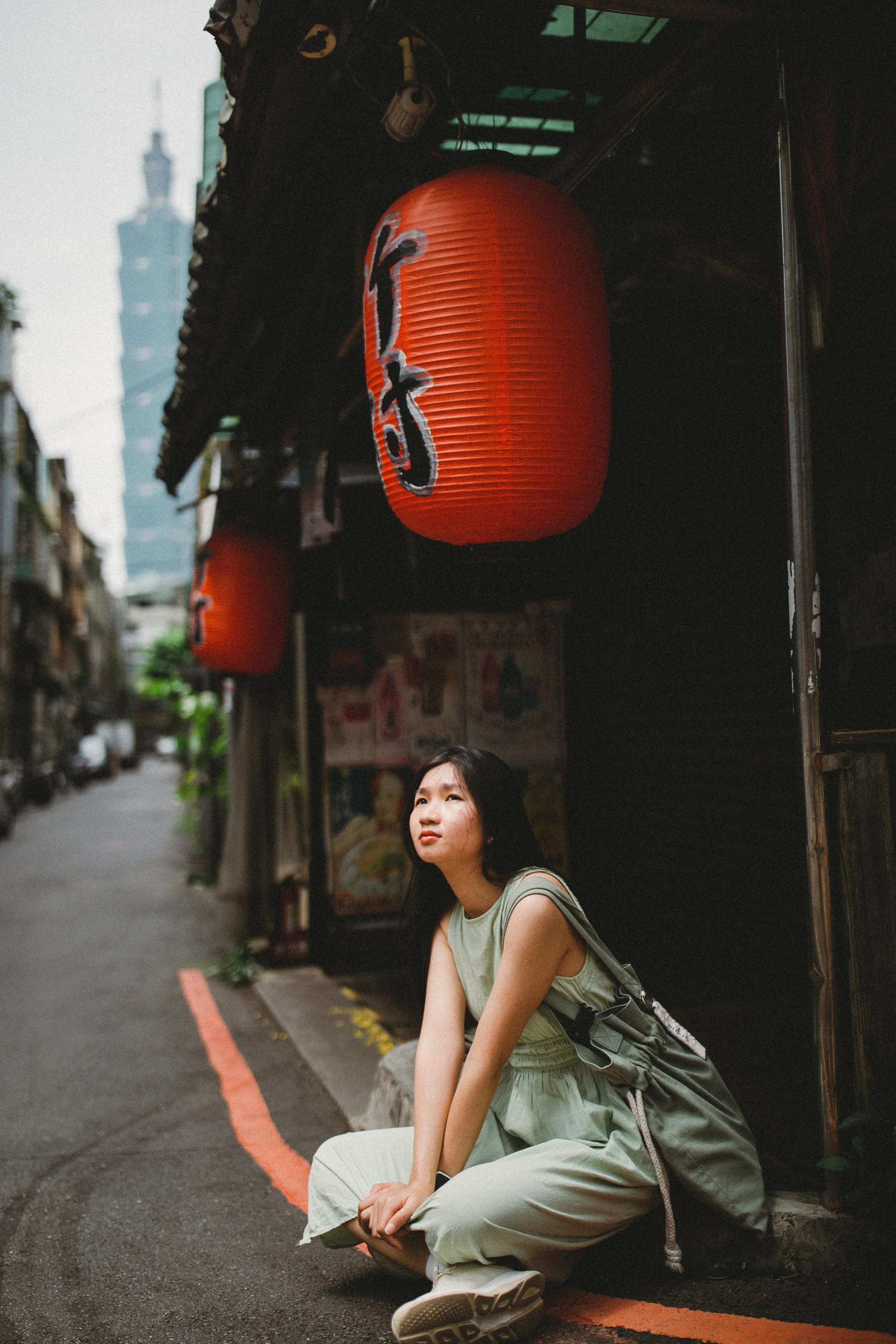 A young woman in a light green outfit sitting on the street curb under red Japanese paper lanterns with Japanese characters, in an urban alley with buildings and a mountain in the background.