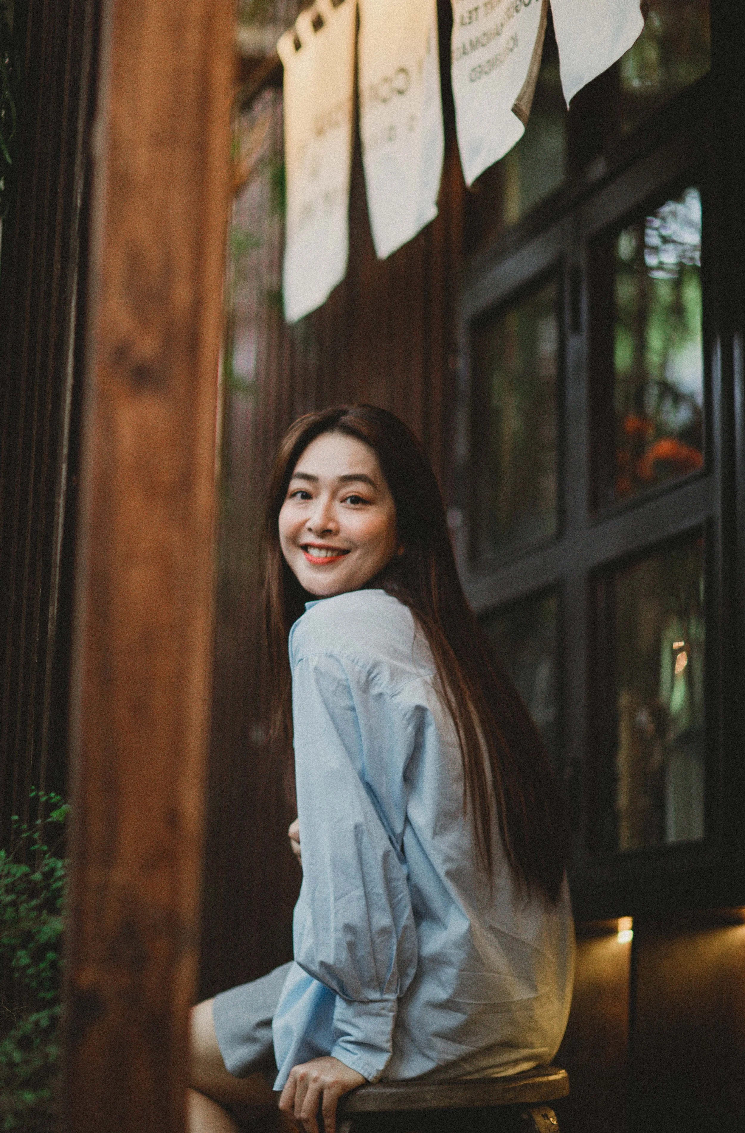 A woman with long dark hair wearing a light blue shirt, sitting on a wooden stool, smiling and looking at the camera, in a cozy, rustic setting with wooden walls and decorative papers hanging above.