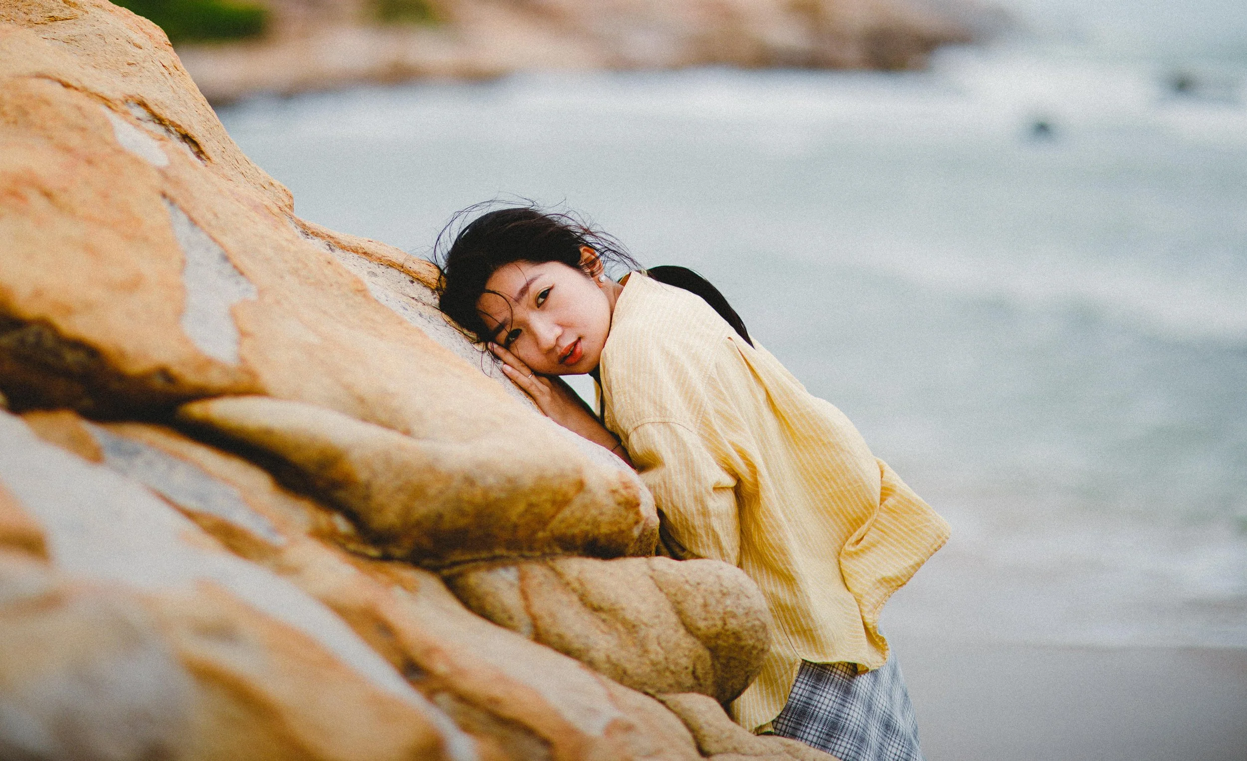 Young woman with black hair leaning on large sandstone rocks at the beach, wearing a yellow striped shirt and grey plaid shorts, with waves in the background.