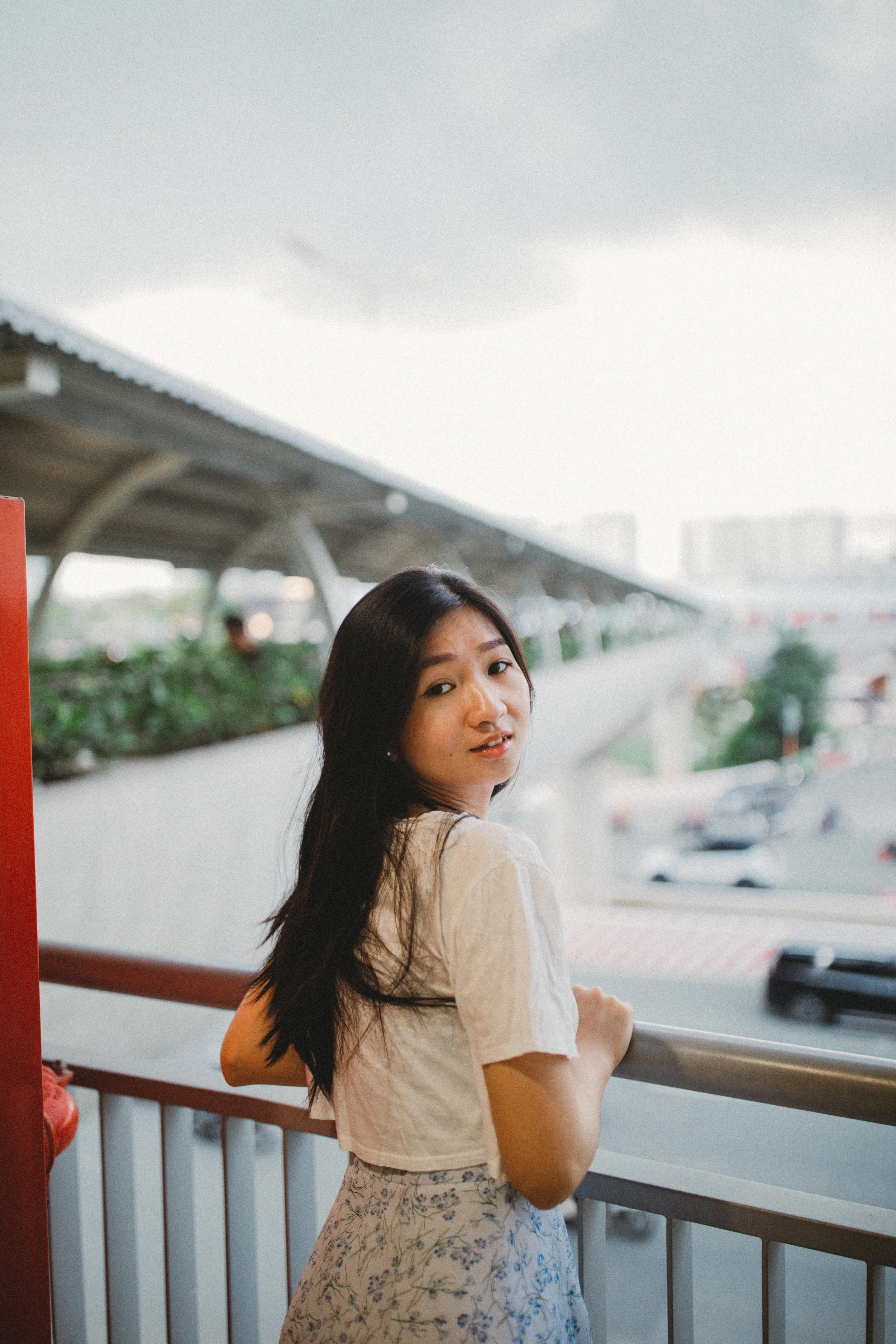A young woman with long dark hair standing on an outdoor observation deck, looking over a busy urban street and parking lot during daytime.