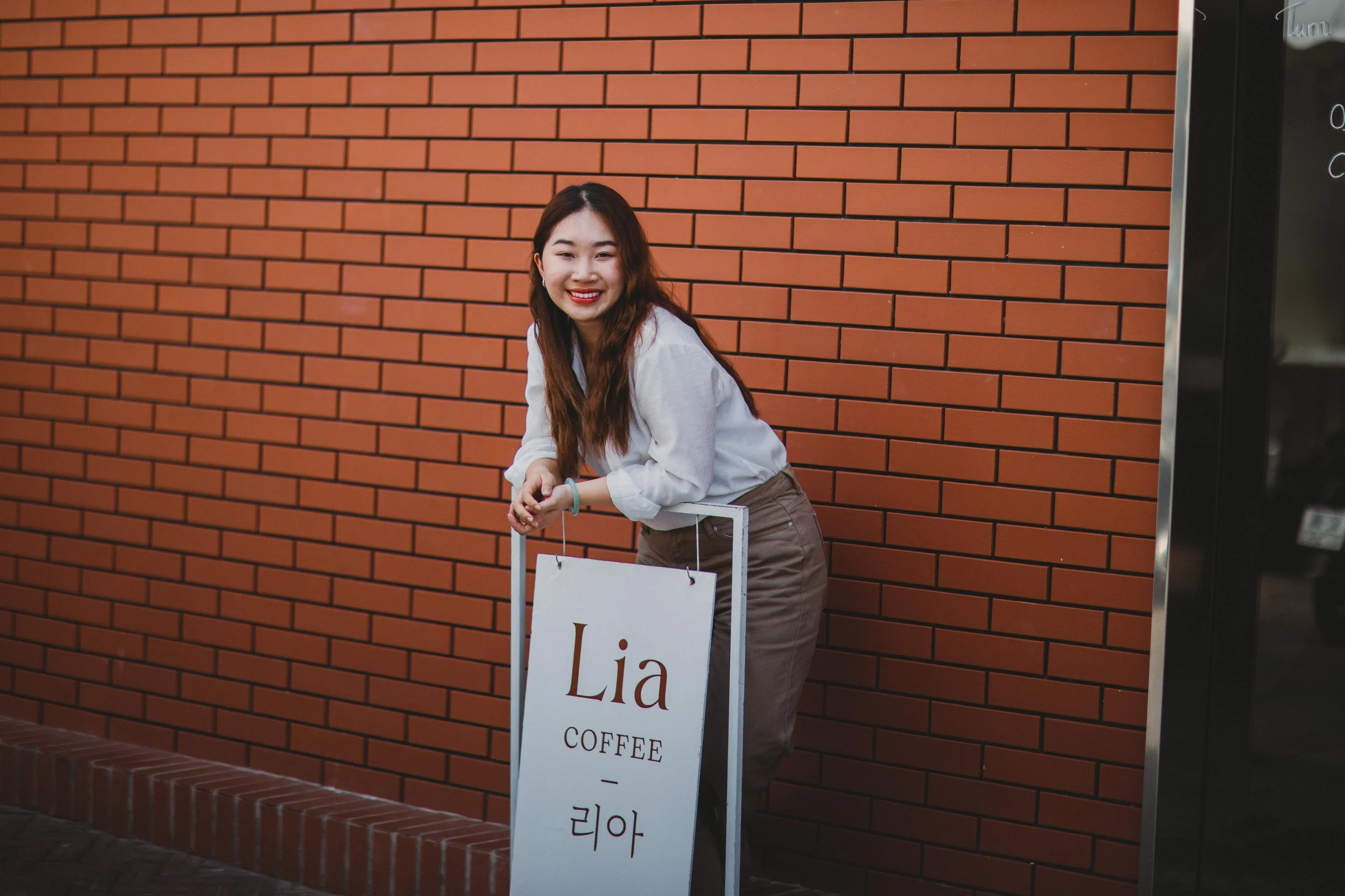 A young woman with long brown hair wearing a white shirt and brown pants, smiling and leaning on a white sign that reads "Lia Coffee" with Korean characters below, standing against a red brick wall.