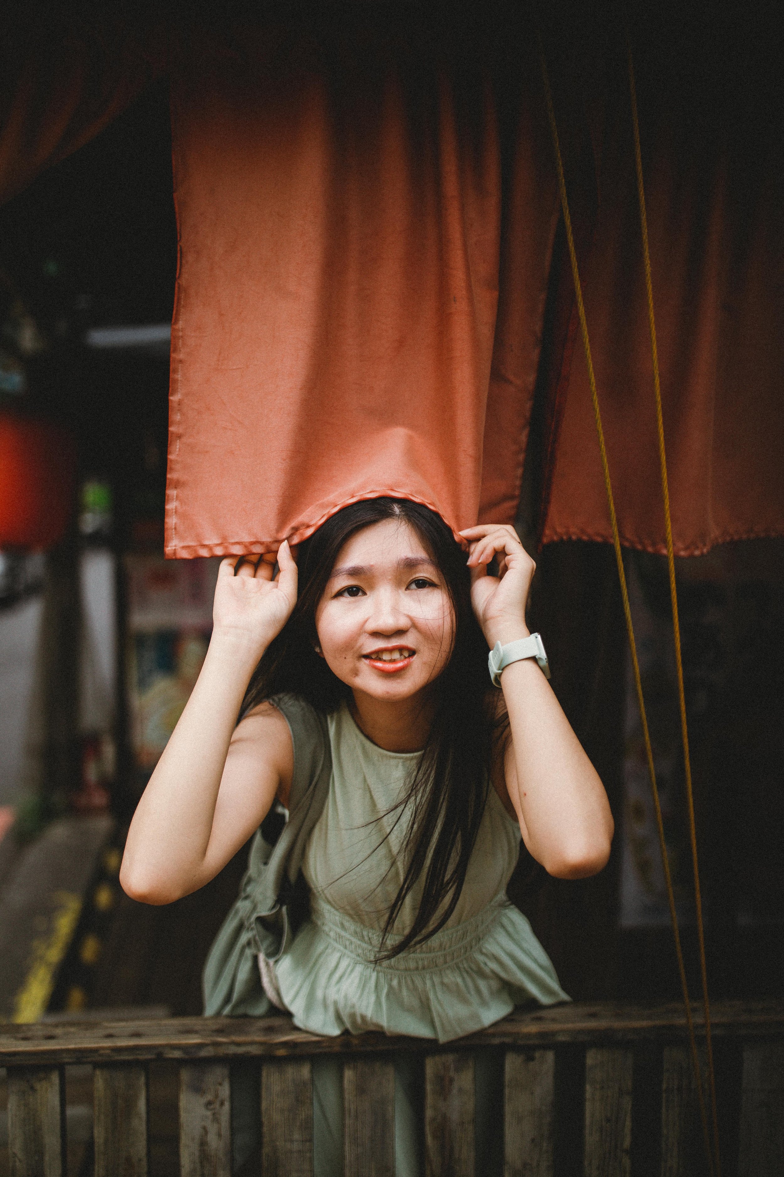Young woman peeking through a red curtain, holding it open with both hands, outdoors with blurred background.
