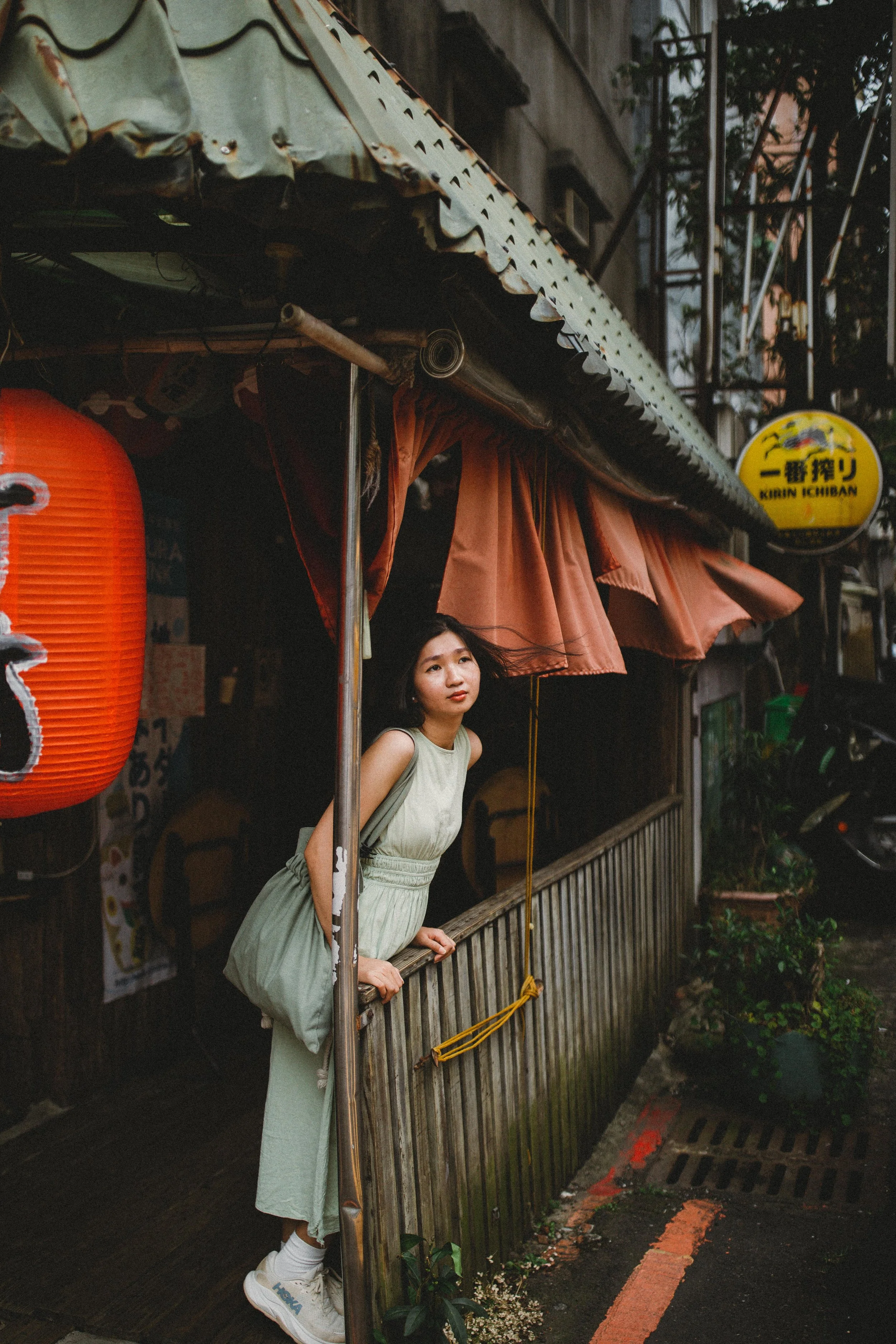 A young woman in a light green dress leaning on a wooden railing outside a building with a yellow sign and red paper lanterns, under a dark canopy and surrounded by urban elements.
